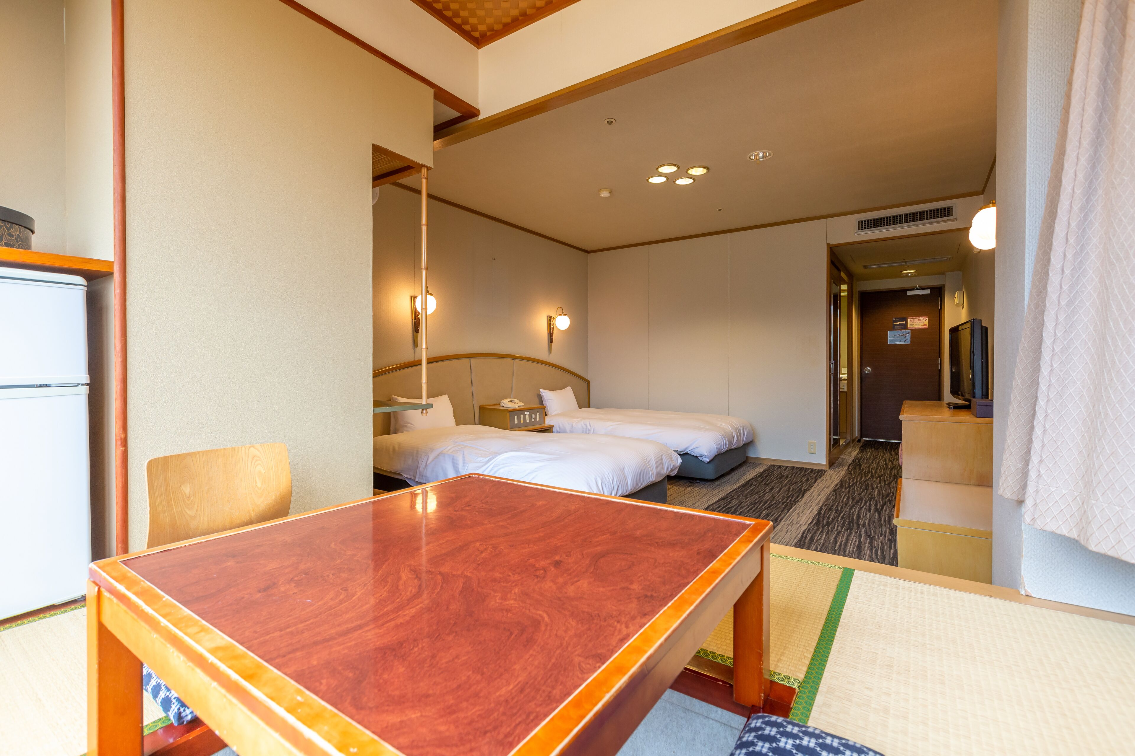 A hotel room with a wooden table, two beds, a chair, and a small kitchenette.