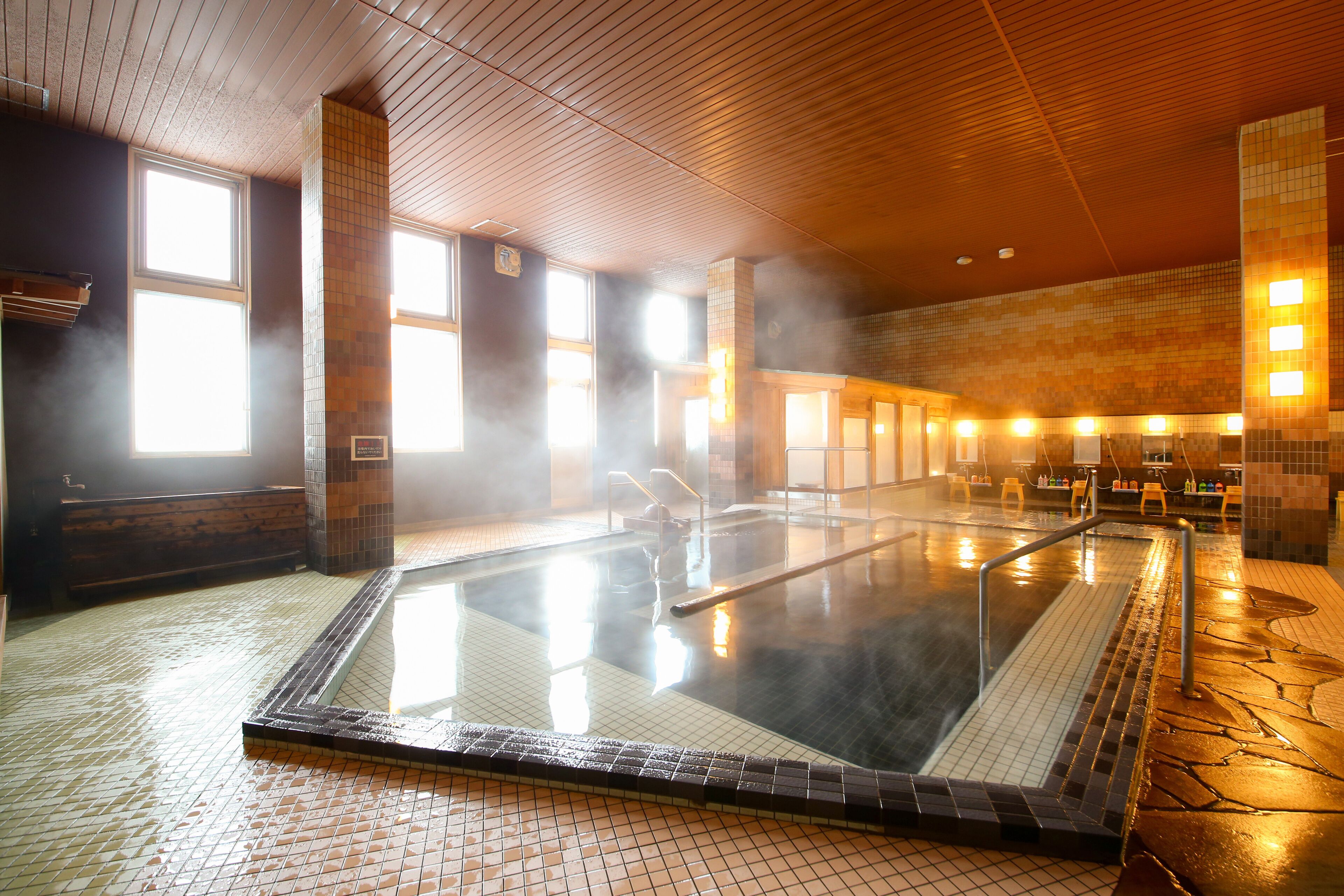 A large indoor swimming pool with tiled walls and a wooden ceiling, surrounded by benches and a tiled floor.