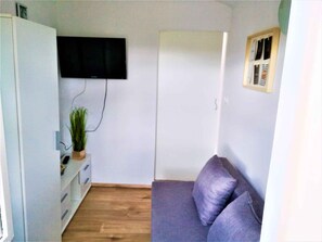 Una stanza piccola e arredata in modo essenziale, con pavimento in legno, un armadio bianco, una TV a parete, un divano viola e una piccola pianta.