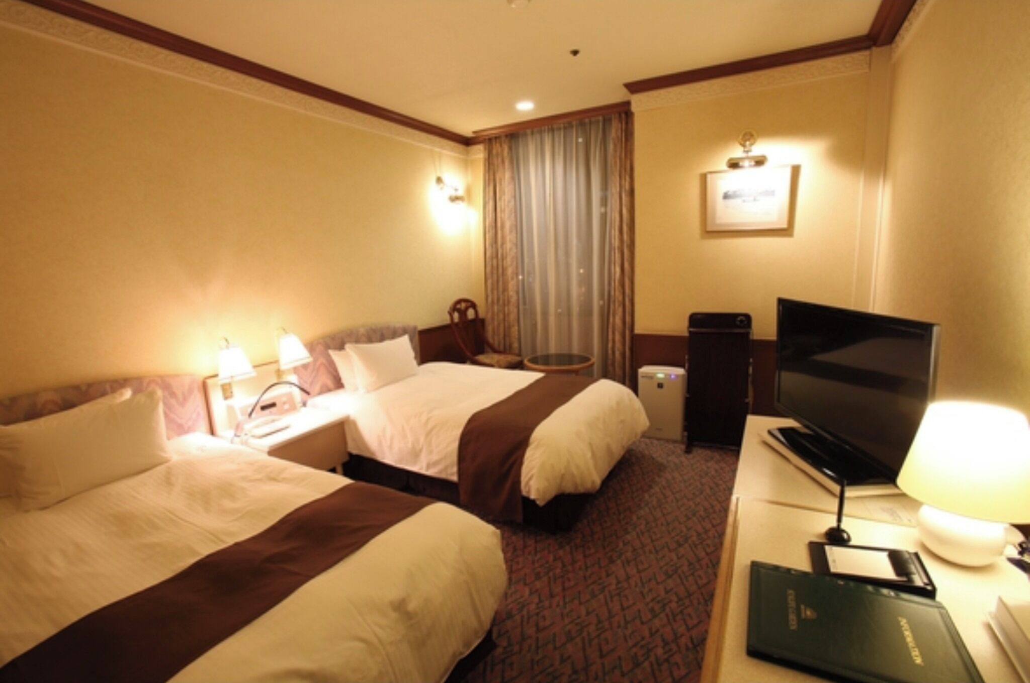 Ein Hotelzimmer mit zwei Betten, einem Fernseher, einem Schreibtisch mit Lampe und einem fenster mit Vorhängen.