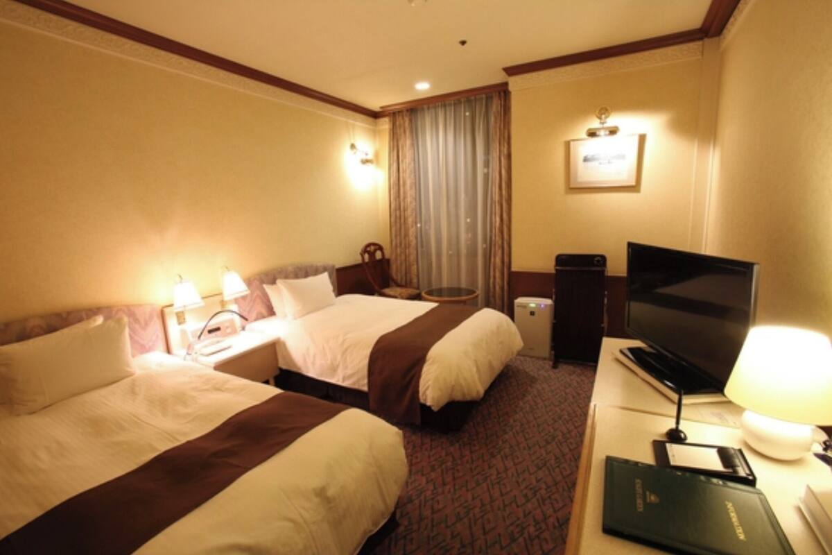 Ein Hotelzimmer mit zwei Betten, einem Fernseher, einem Schreibtisch mit Lampe und einem fenster mit Vorhängen.
