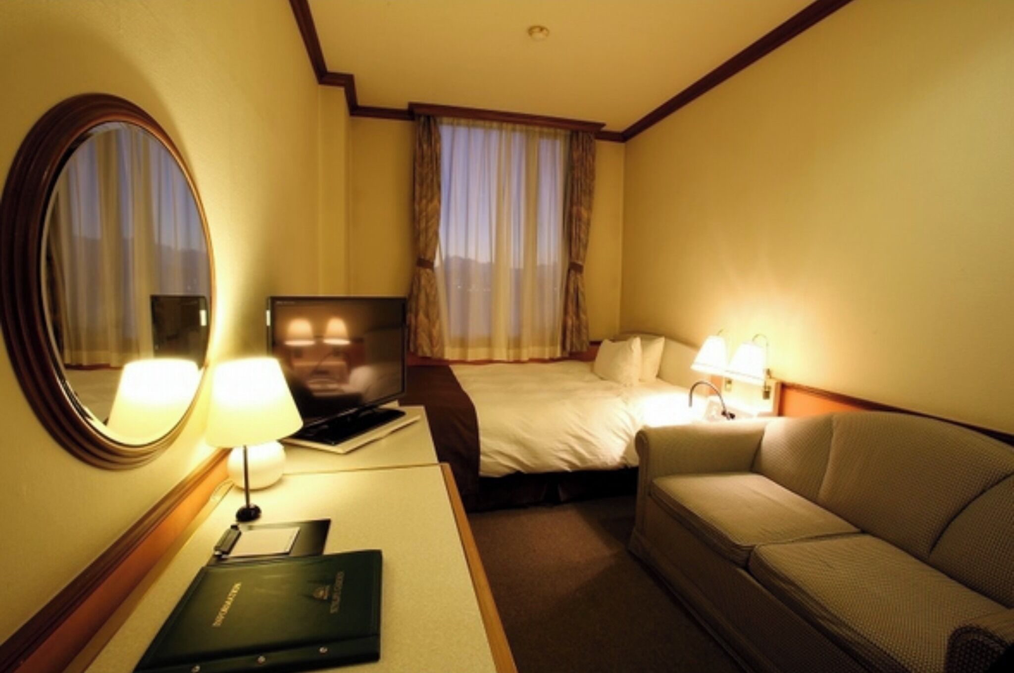 Ein Hotelzimmer mit einem Bett, einem Sofa, einem Schreibtisch mit Lampe, einem Fernseher und einem Spiegel.