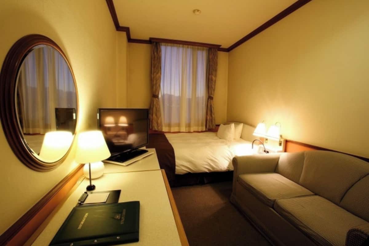 Ein Hotelzimmer mit einem Bett, einem Sofa, einem Schreibtisch mit Lampe, einem Fernseher und einem Spiegel.