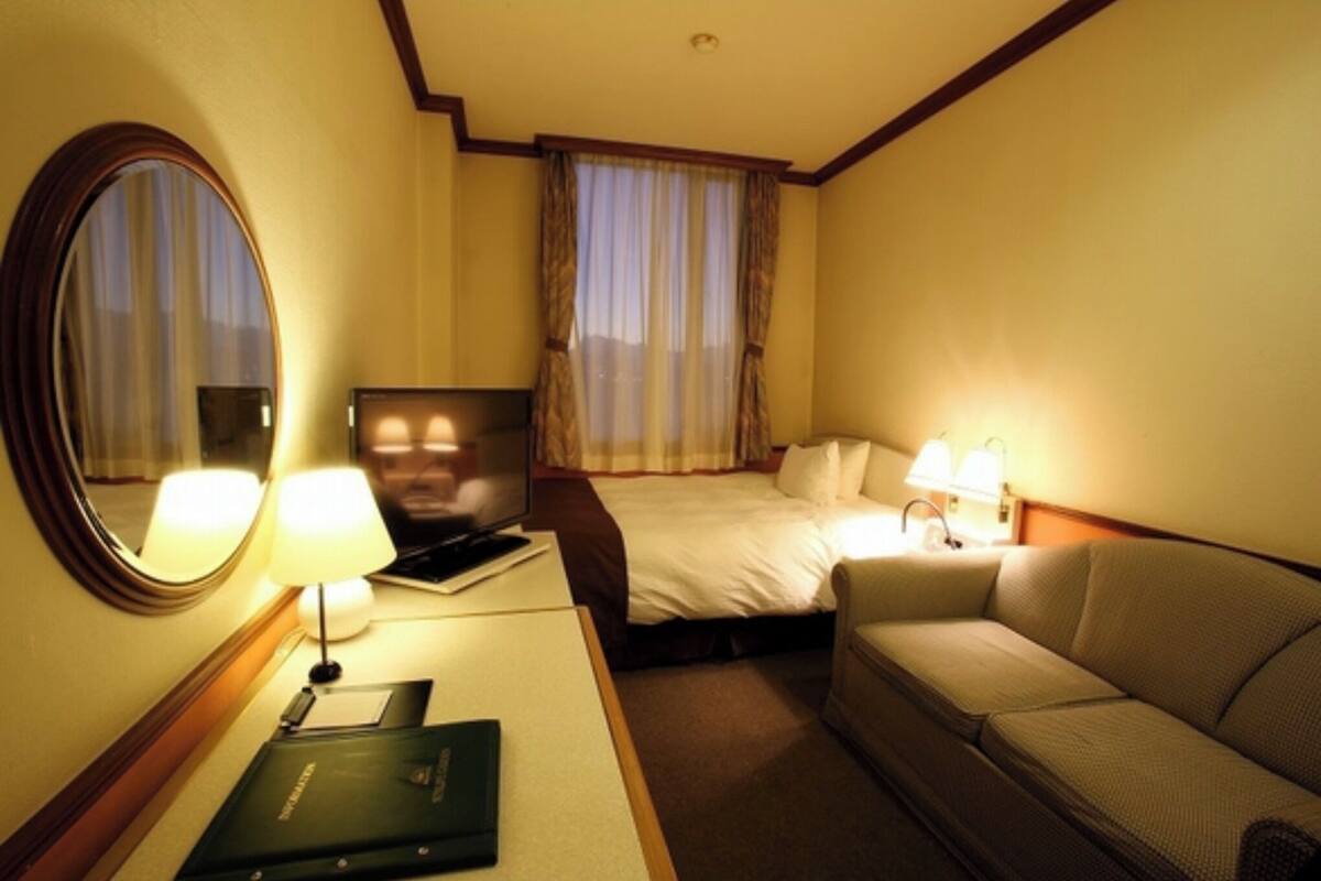 Ein Hotelzimmer mit einem Bett, einem Sofa, einem Schreibtisch mit Lampe, einem Fernseher und einem Spiegel.