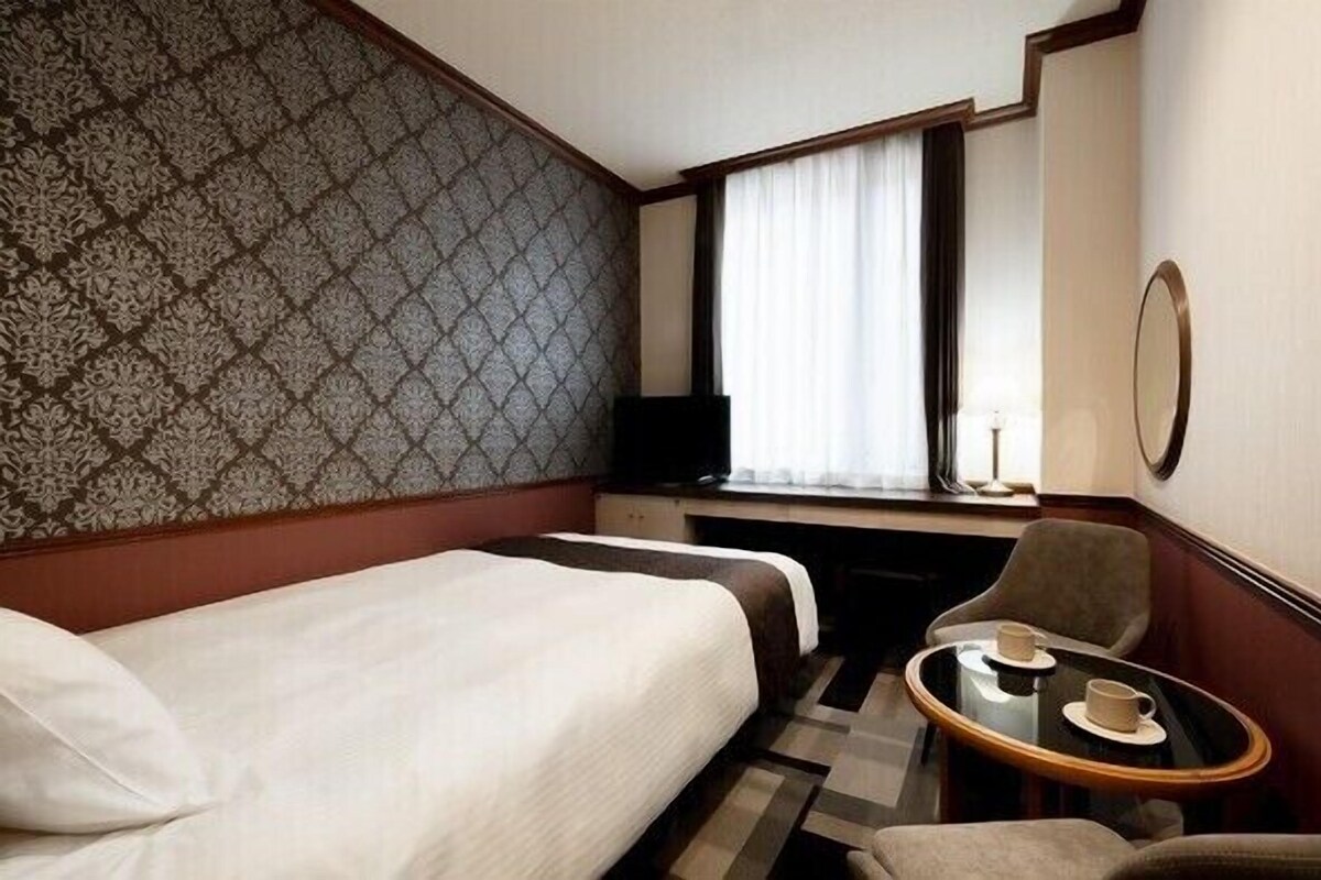 Hotelkamer met een bed, een bureau, een stoel, een klein tafeltje met bekers en een raam met gordijnen.