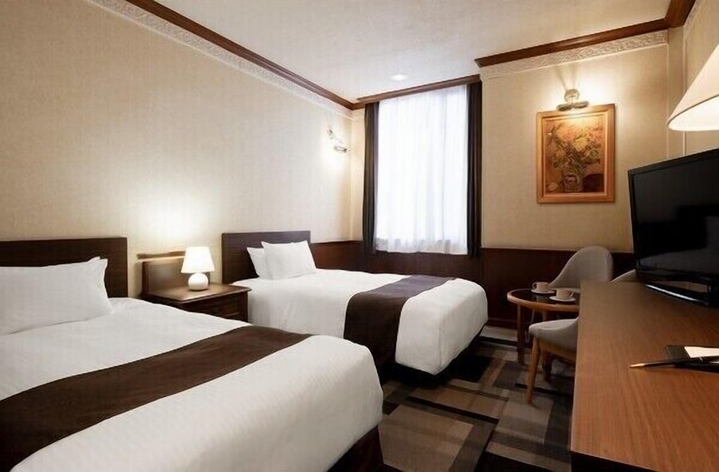 Ein Hotelzimmer mit zwei Betten, einem Schreibtisch, einem Fernseher und einem Fenster mit Vorhängen.