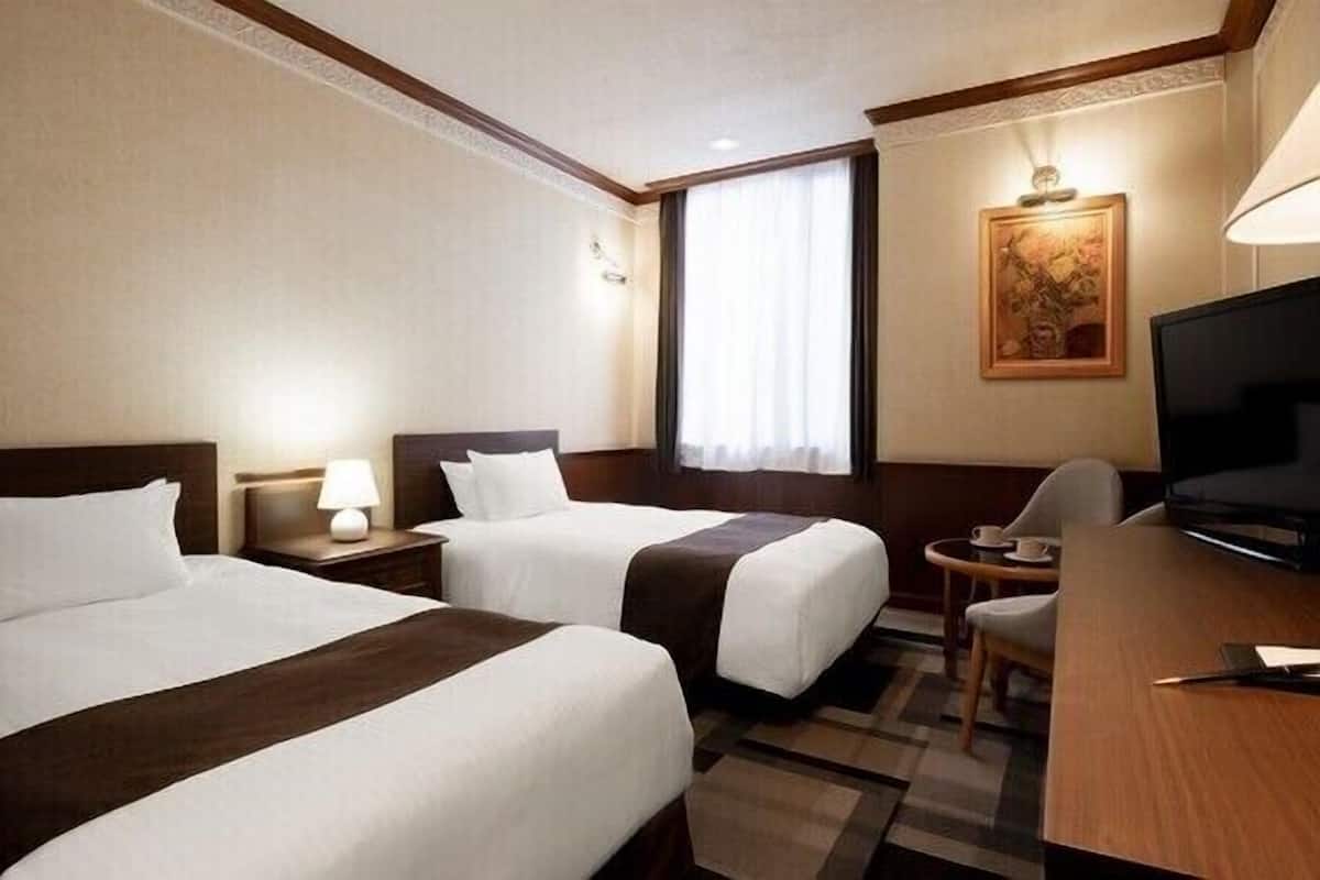 Ein Hotelzimmer mit zwei Betten, einem Schreibtisch, einem Fernseher und einem Fenster mit Vorhängen.