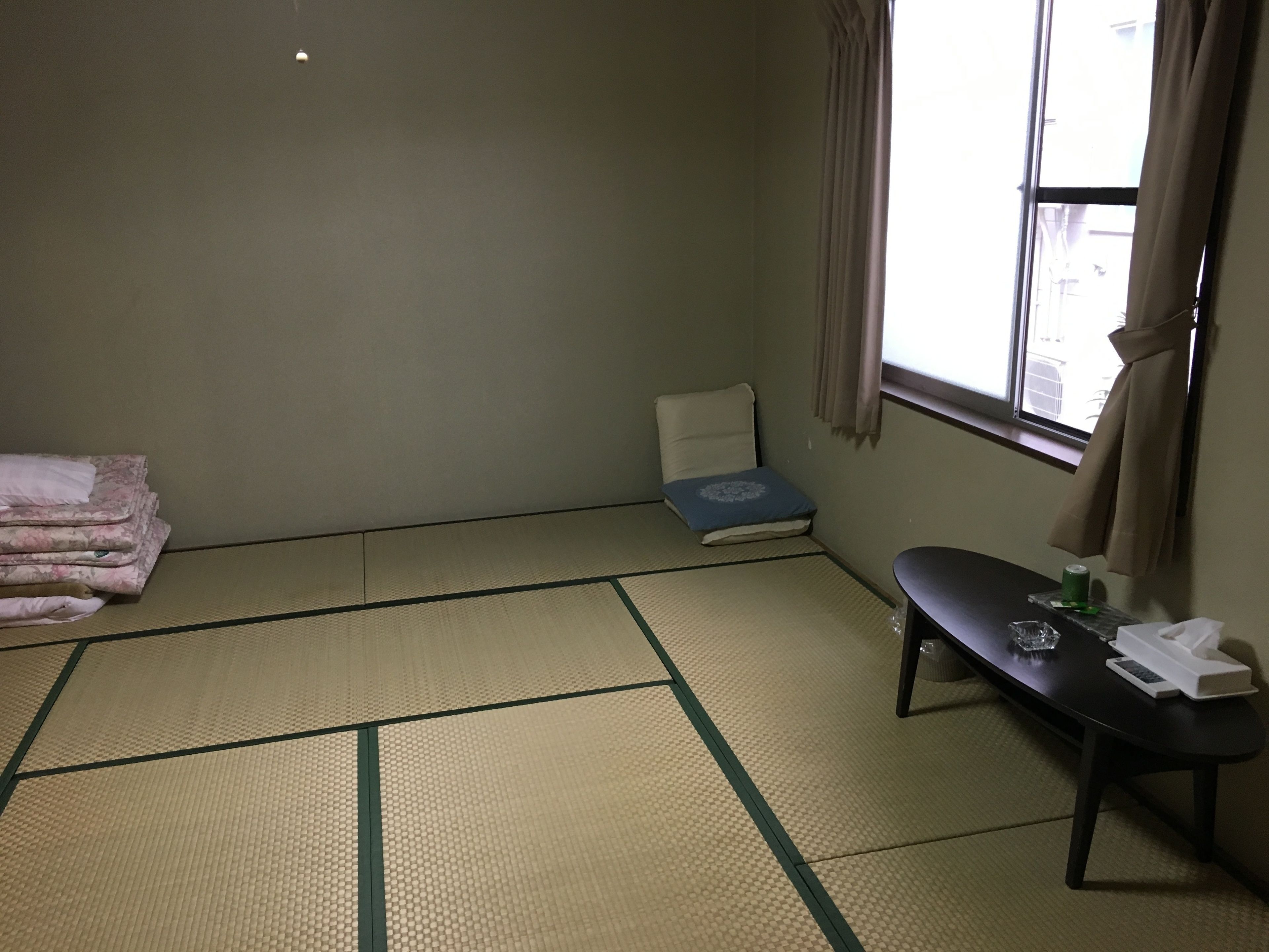 A room with tatami mat flooring, a bed with a blue pillow, a small table with a bottle and glasses, and a window with curtains.