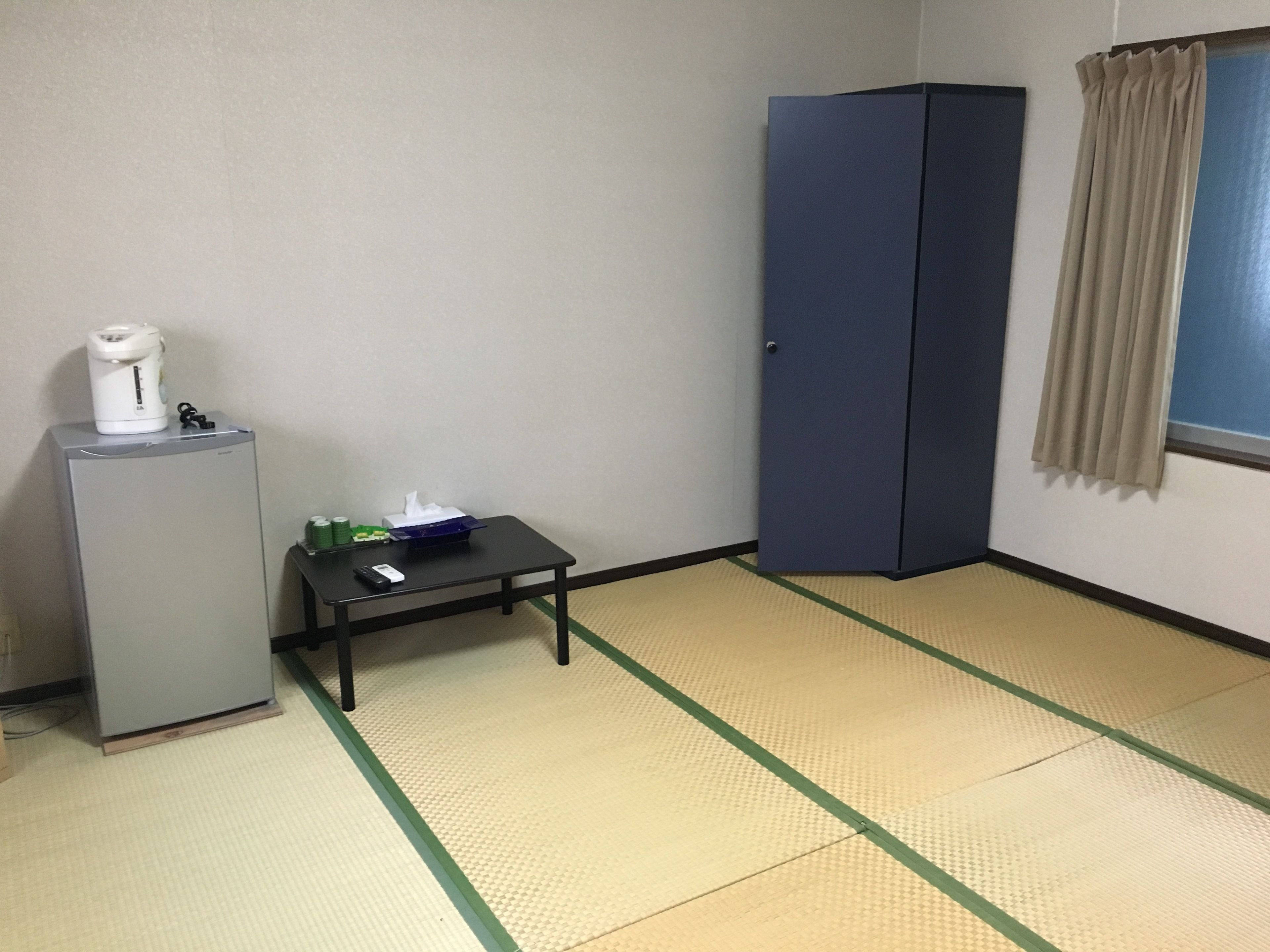 Una stanza con pavimento in tatami, un tavolino con una scatola di fazzoletti e il telecomando, un armadietto e una finestra con tende.