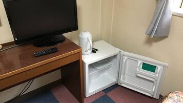 A hotel room with a wooden desk, a black TV, a white kettle, and a mini-fridge.