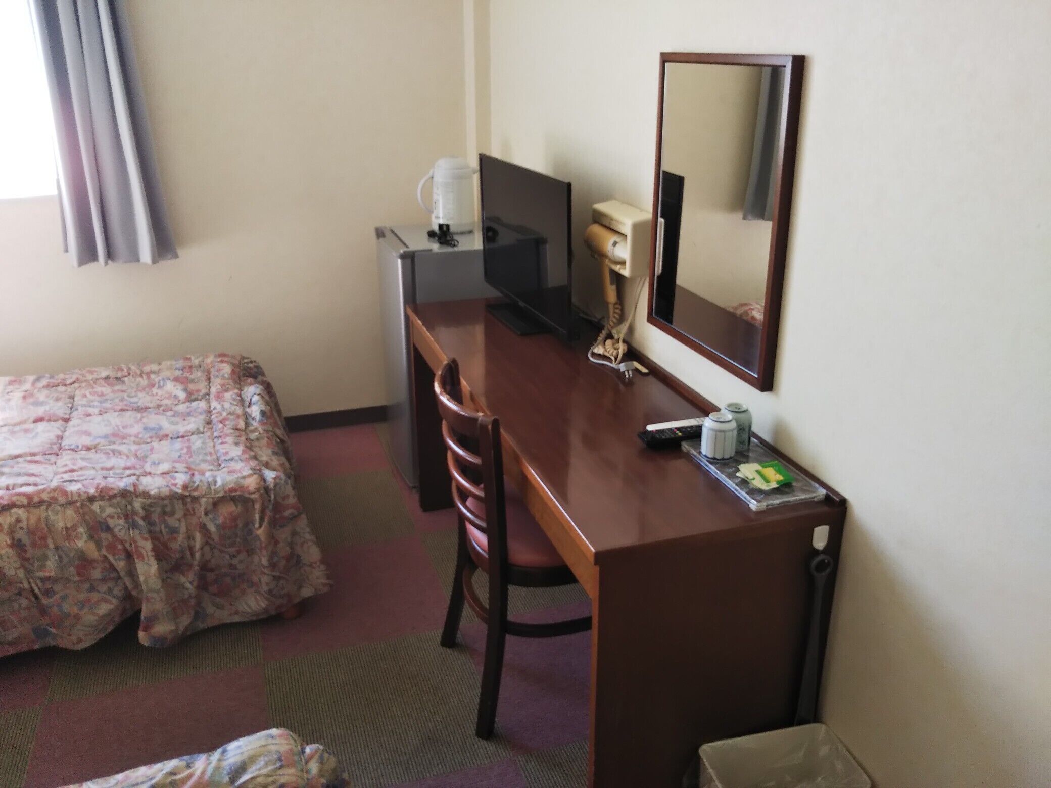 Ein Hotelzimmer mit einem Bett, einem Schreibtisch, einem Stuhl, einem Fernseher, einem Spiegel und einem kleinen Tisch mit Fernbedienung und einer Dose.