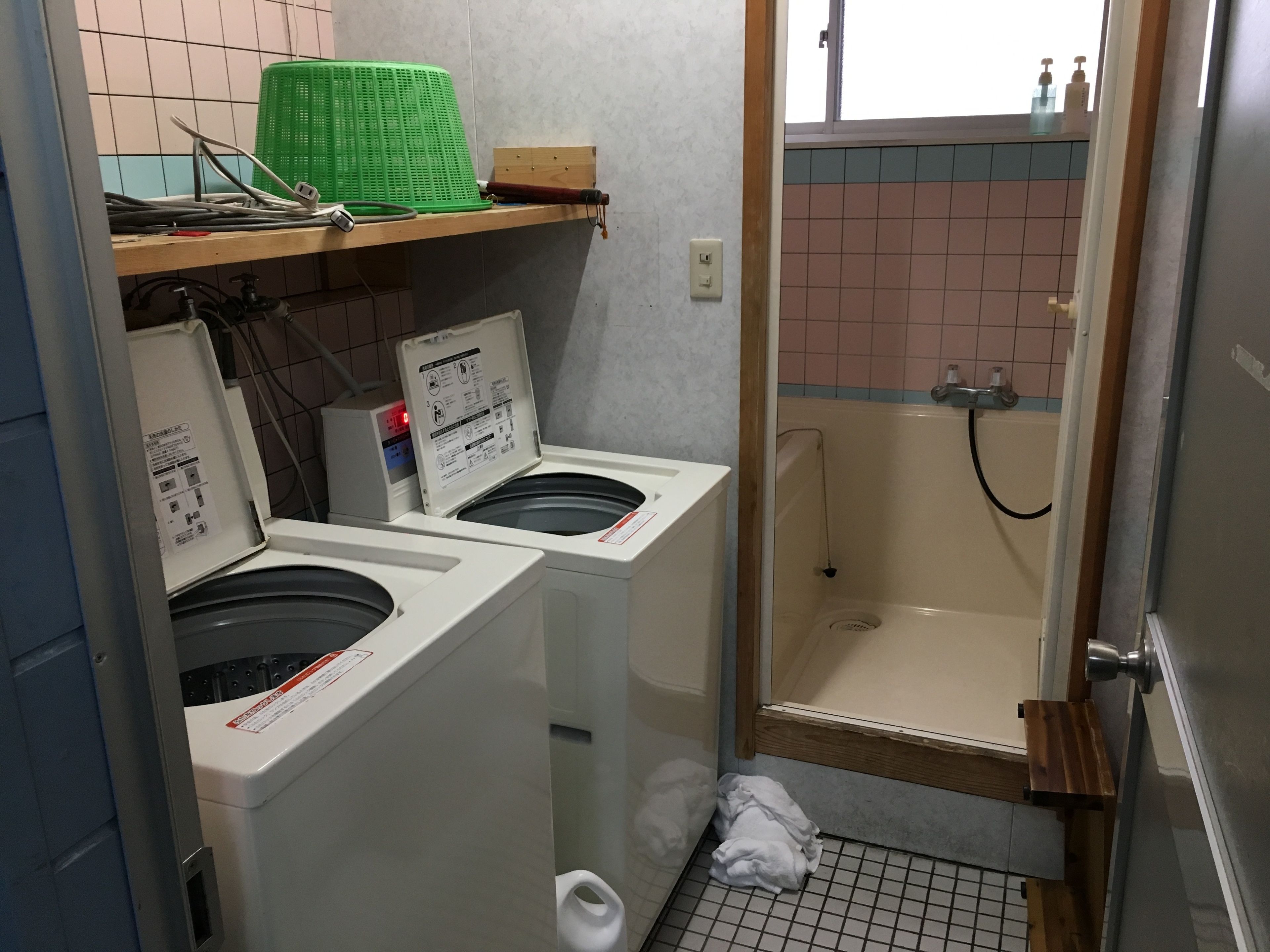 Un lavadero pequeño con dos lavadoras, una zona de ducha y un estante de madera con diversos objetos.