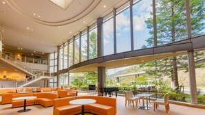 Un hall d’entrée spacieux, doté de sièges orange, d’un piano et de grandes fenêtres donnant sur un paysage verdoyant.