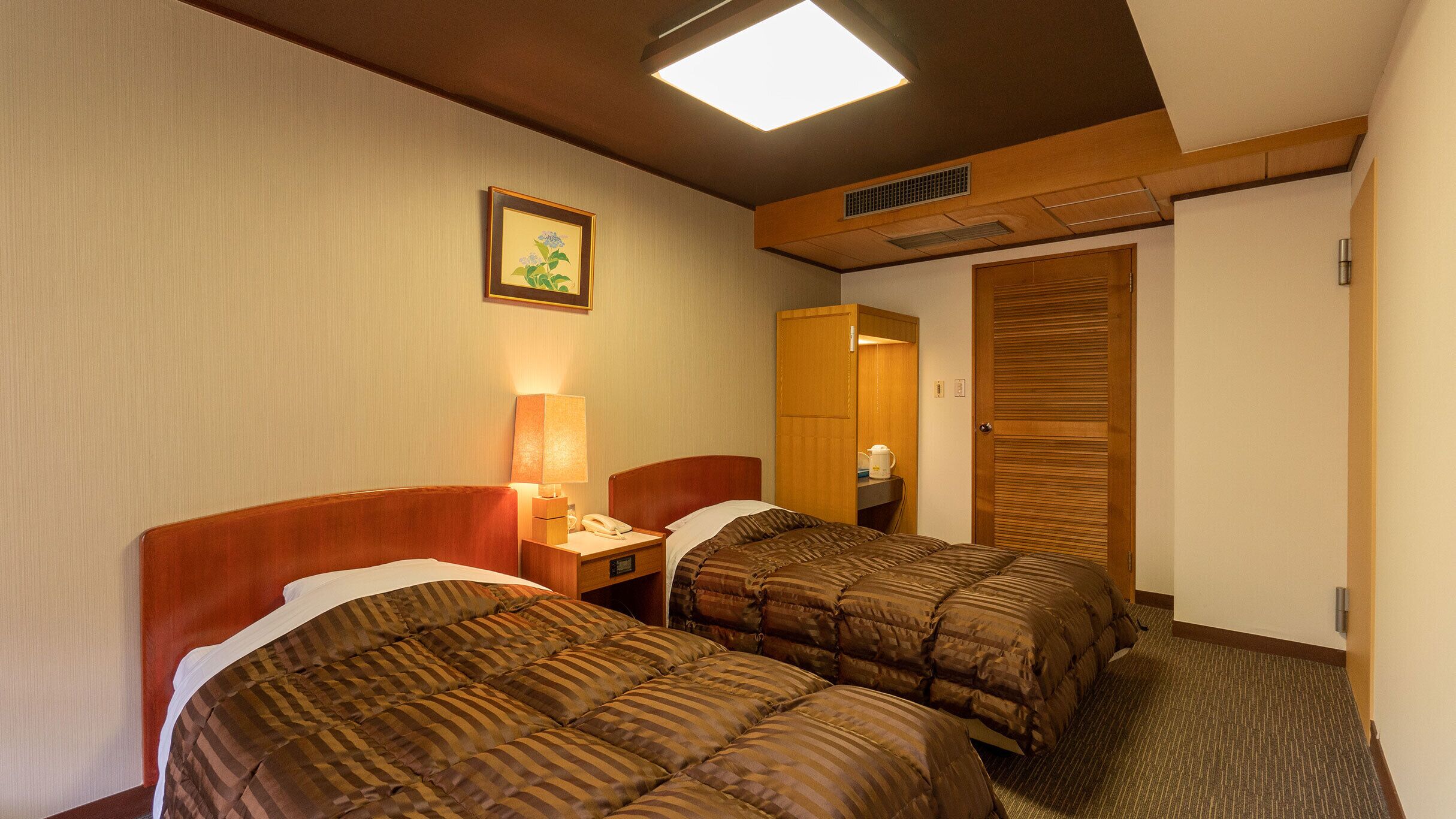 A hotel room with two beds, a bedside table with a lamp, a framed picture on the wall, and a wardrobe.