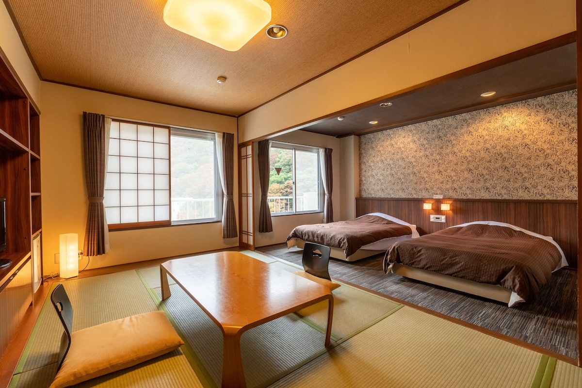 Een traditionele Japanse kamer met tatami vloeren, een lage tafel, twee bedden en uitzicht op groen buiten.