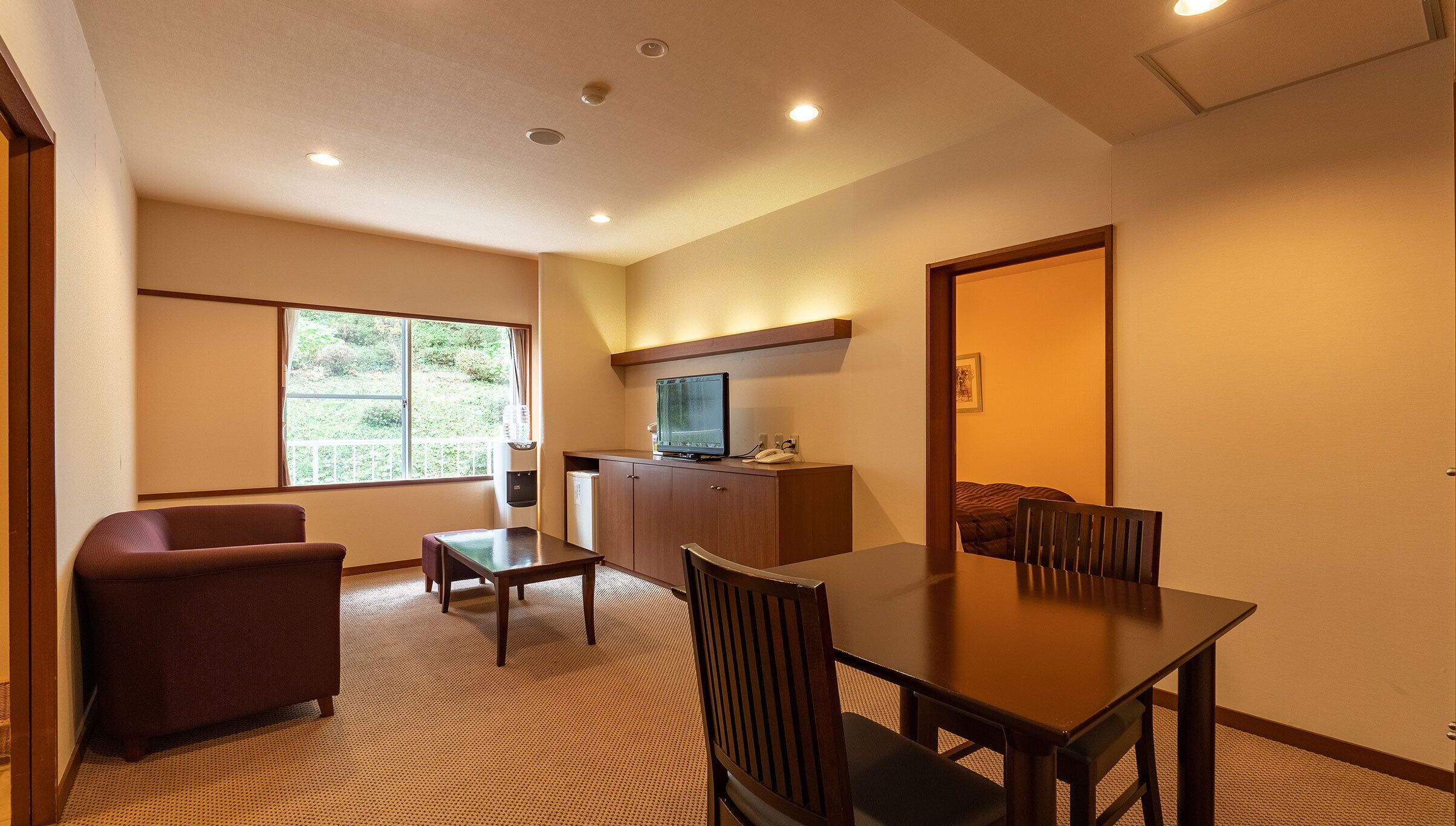 A hotel room with a dining area, a sofa, a TV on a cabinet, and a large window.