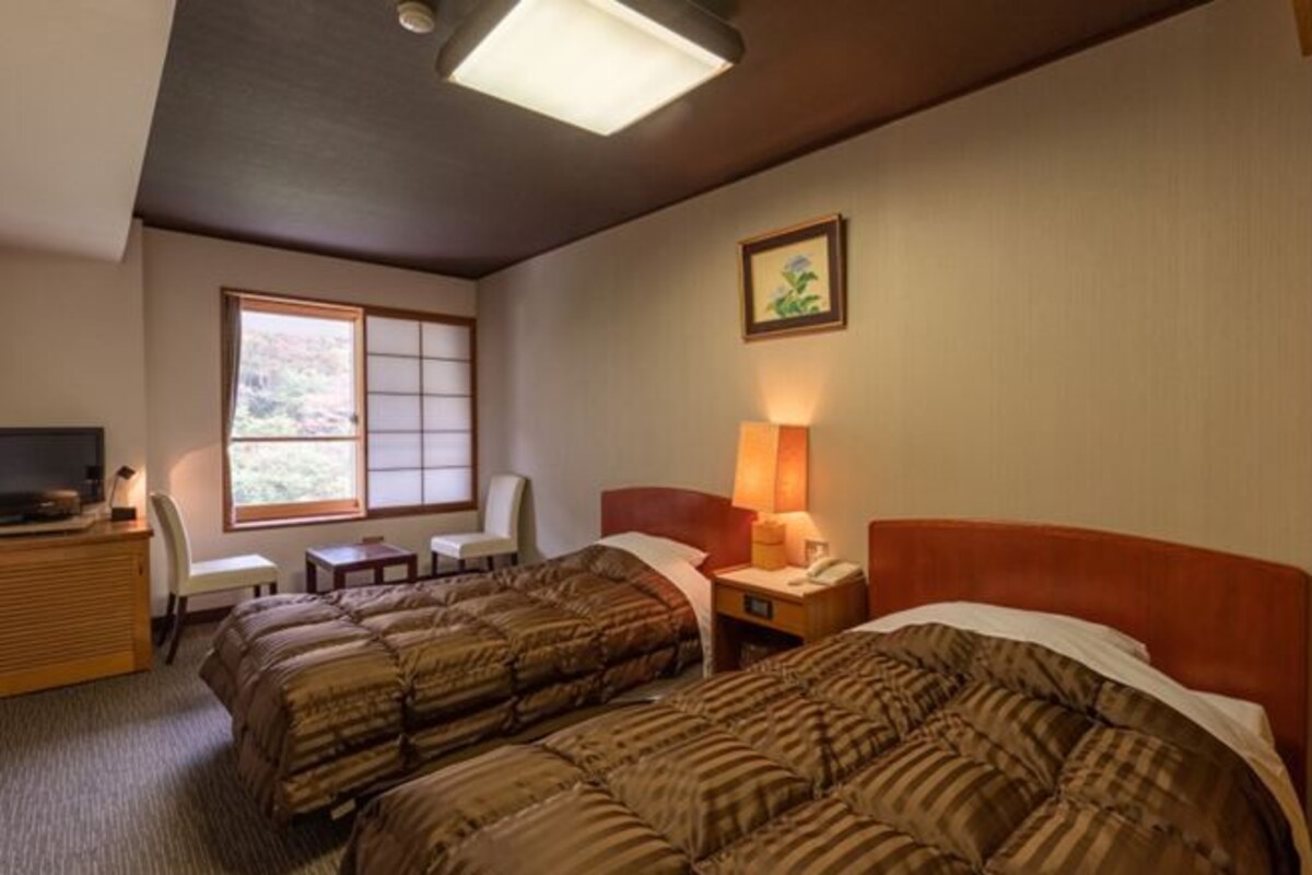 A hotel room with two beds, a desk, a chair, a window, and a painting on the wall.