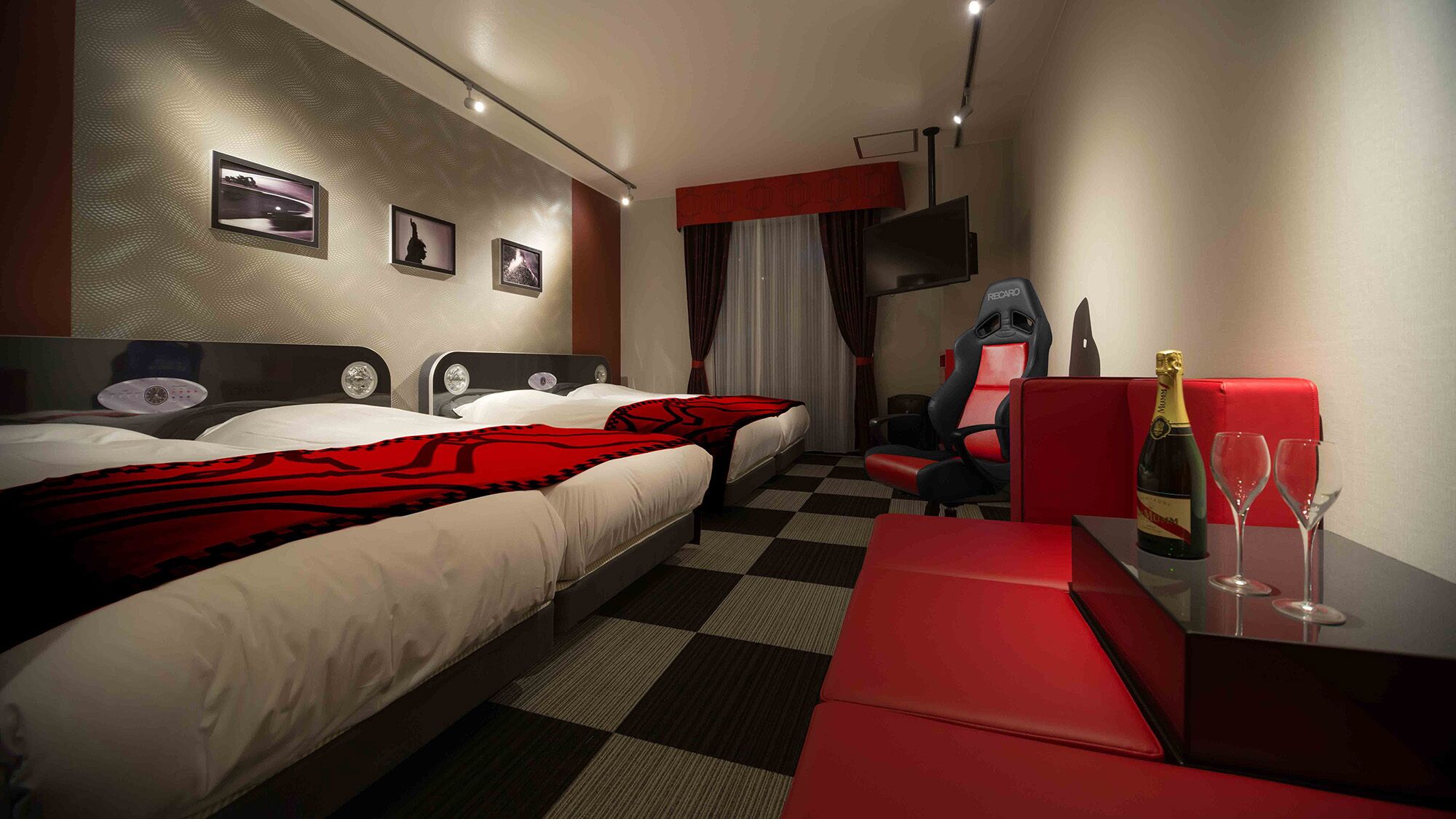 Habitación de hotel con dos camas, televisión, una silla roja, una mesa con una botella y vasos, y obras de arte en la pared.