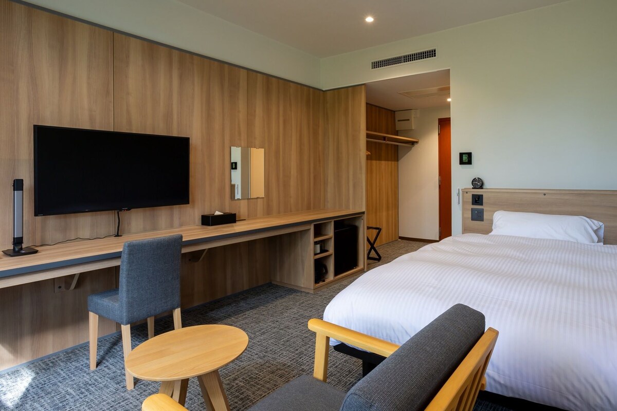 A hotel room with a bed, a desk with a chair, a TV, and a wardrobe.