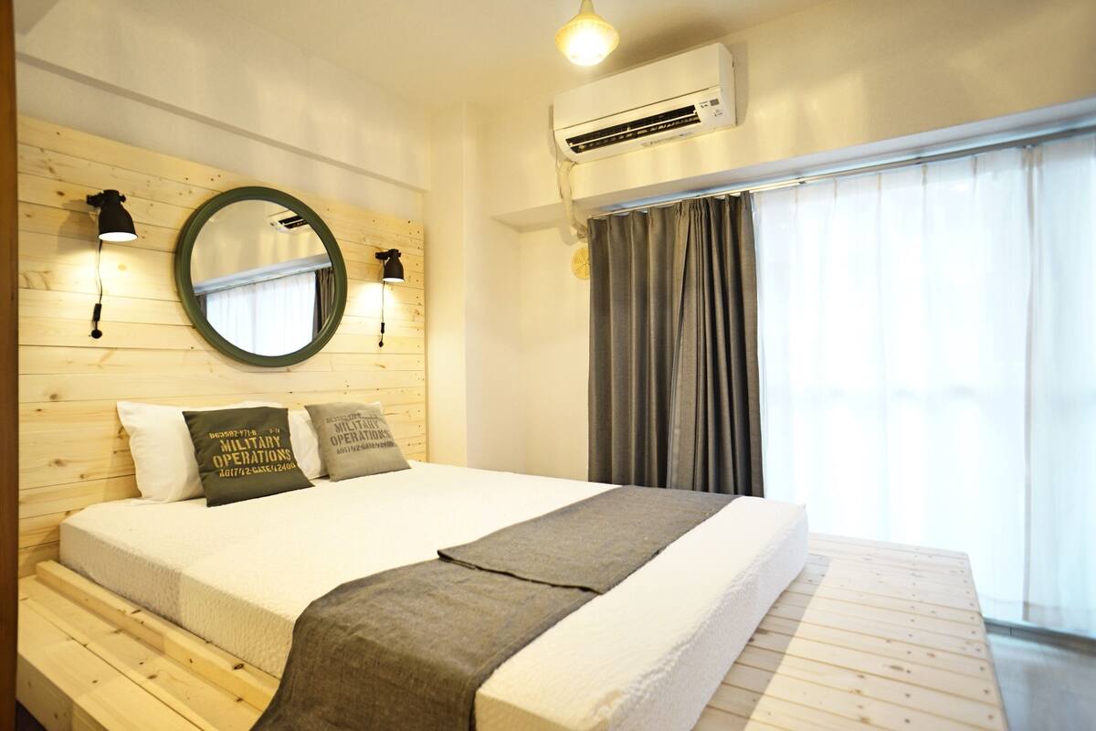 Habitación de hotel con cabecera de madera, cama grande con colchón blanco y ropa de cama gris, espejo redondo, dos lámparas fijadas a la pared, equipo de aire acondicionado y ventana con cortinas grises.