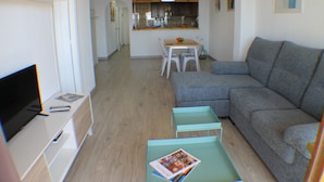 Un soggiorno moderno con un divano a tre posti grigio, un tavolino con una rivista, una TV su un mobile e una cucina sullo sfondo.