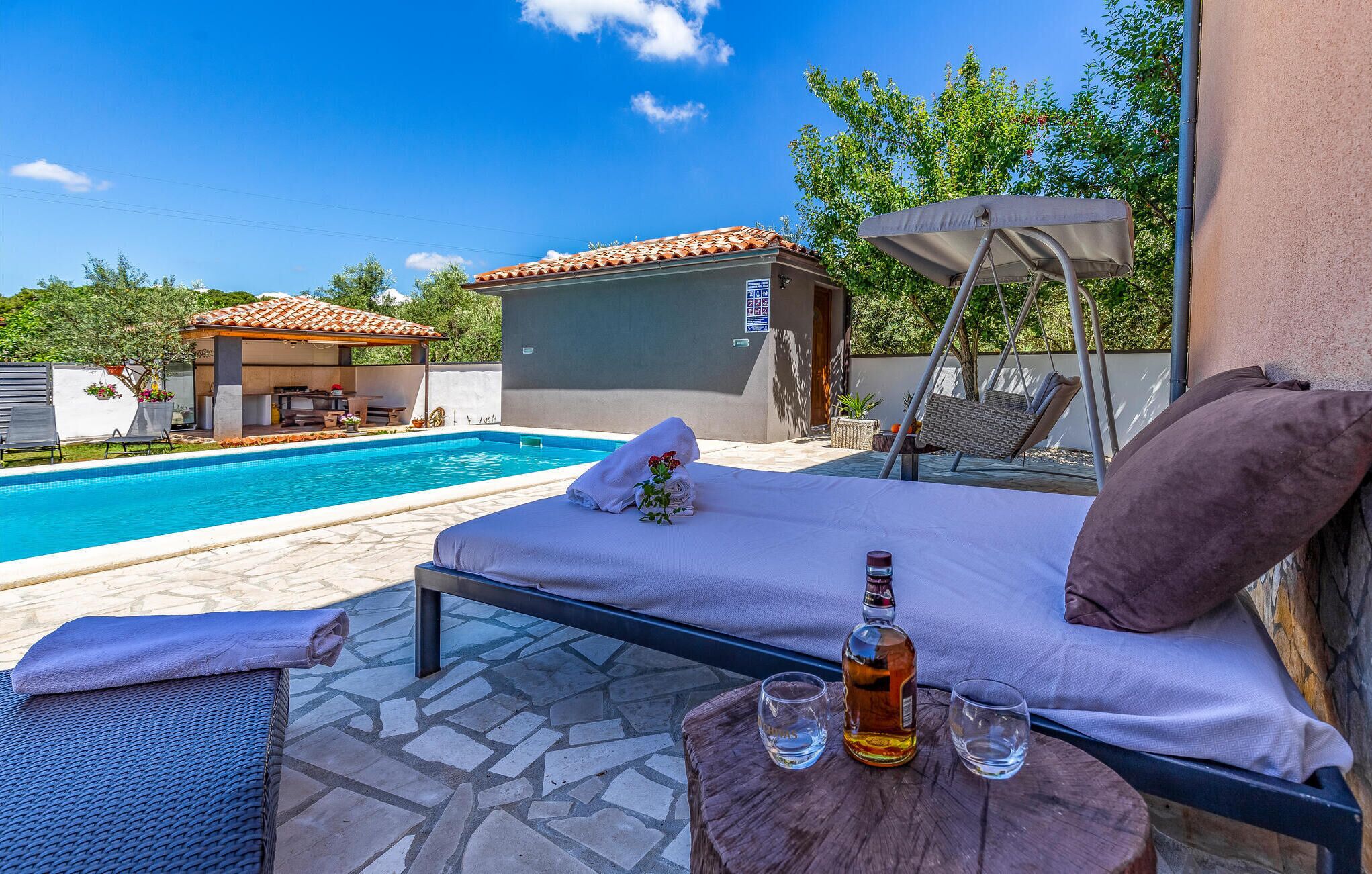A poolside lounge with a bed, a bottle, and glasses on a wooden table.