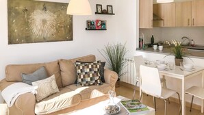 Een woonkamer met een beige bank, een bijzettafeltje met een boek en een glazen vaas, een eethoek met een witte tafel en stoelen, en een keuken met houten kastjes en een hanglamp.