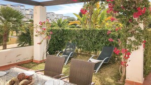 Een terras buiten met rieten meubels, een eettafel en uitzicht op een tuin met diverse planten en bloemen.