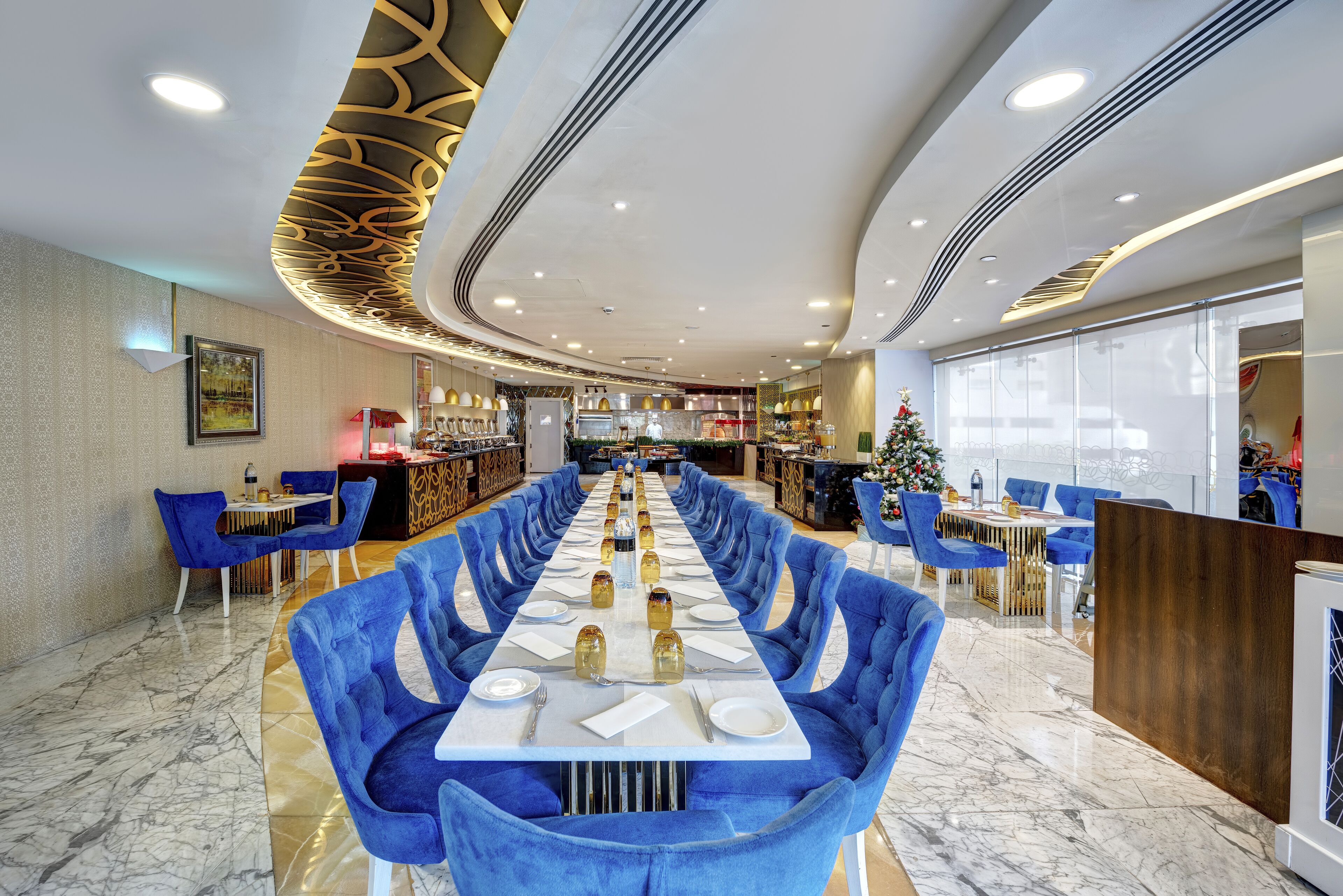 A modern restaurant with a long table set for a meal, blue upholstered chairs, and a decorative ceiling.