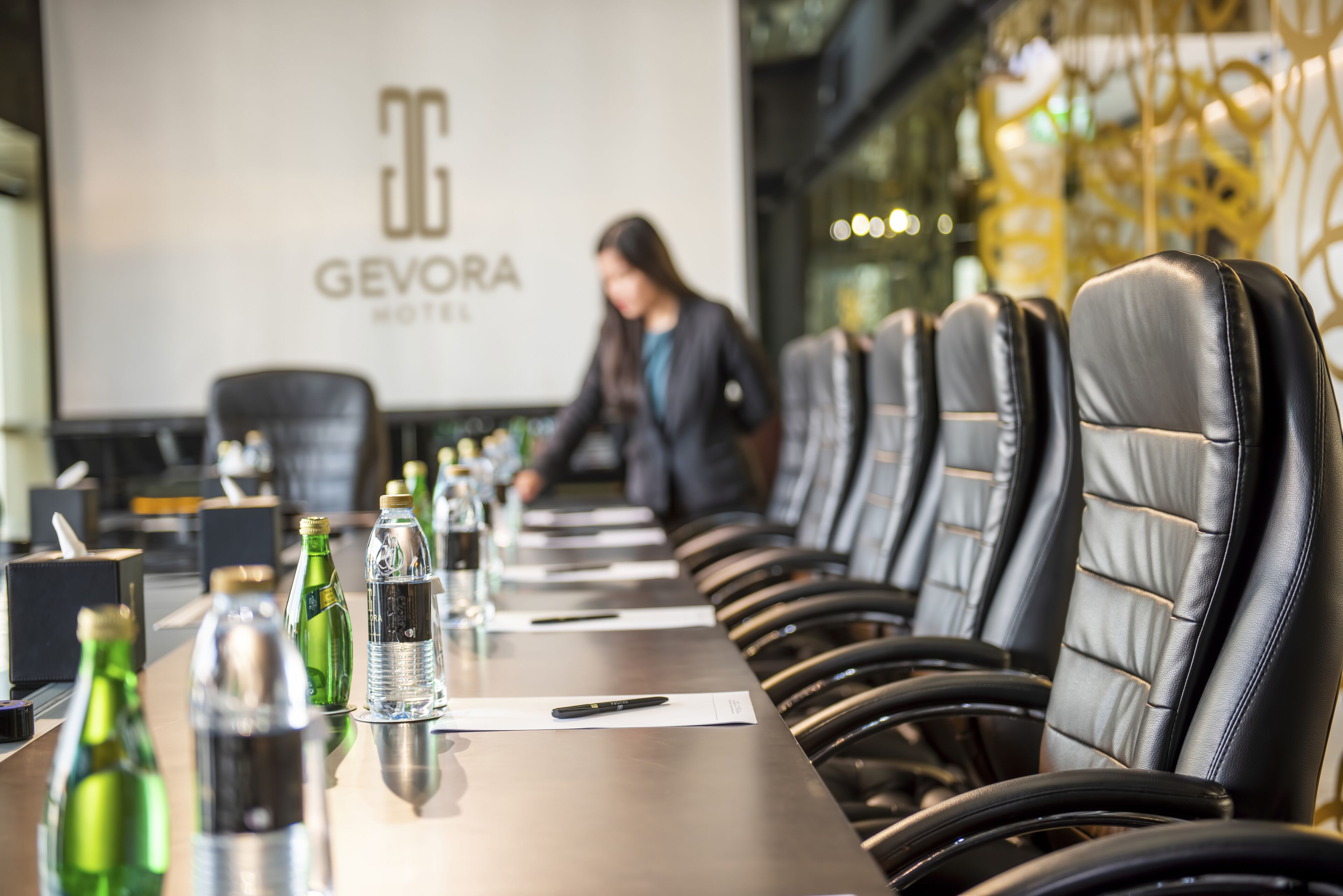 Kokoushuone, jossa on pitkä pöytä, tuoleja ja projektorikangas, jolle on heijastettu teksti ”GEVORA HOTEL”.