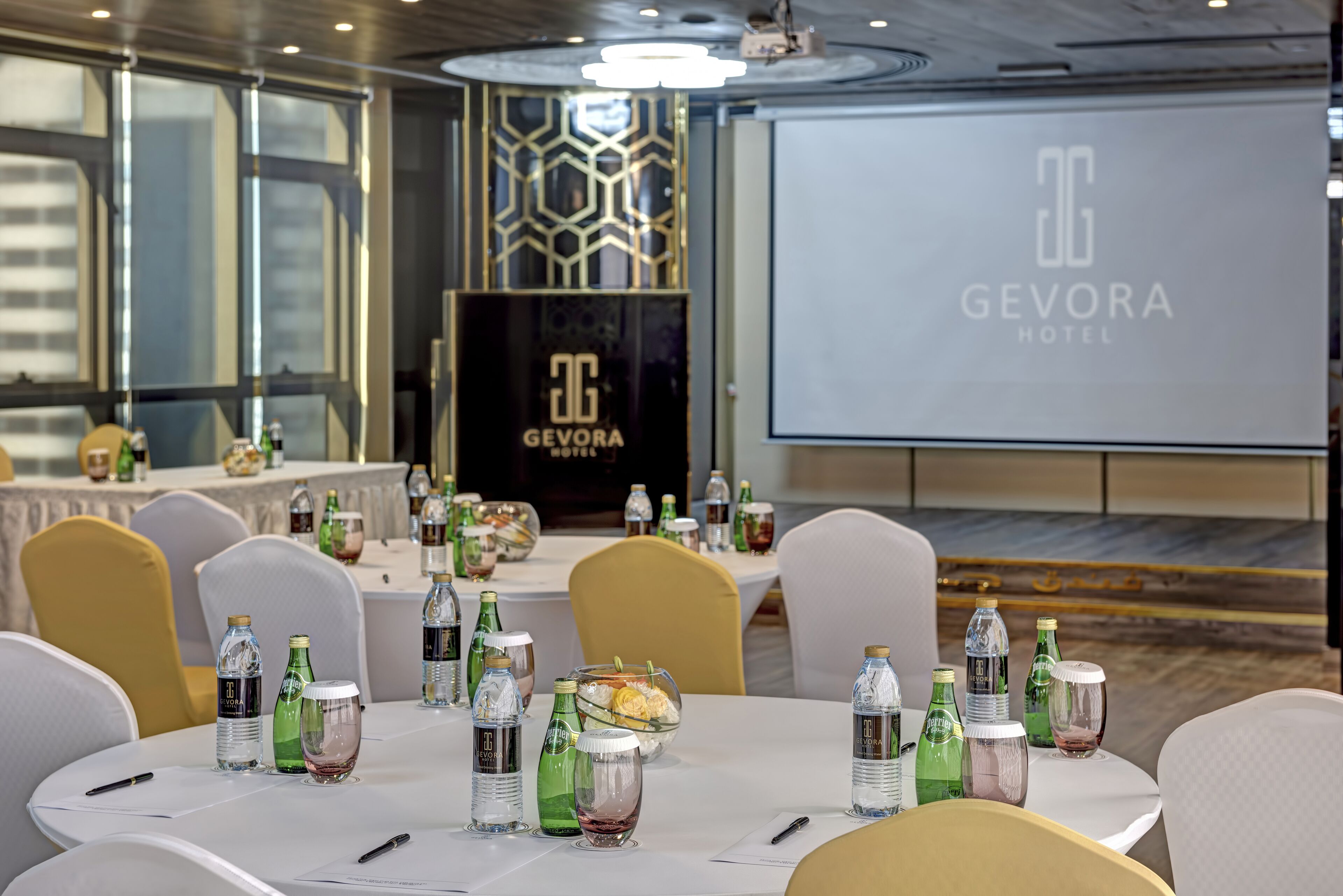Salón de conferencias en el Hotel Gevora con mesas redondas, sillas, botellas de agua y una pantalla proyectora mostrando el nombre del hotel.