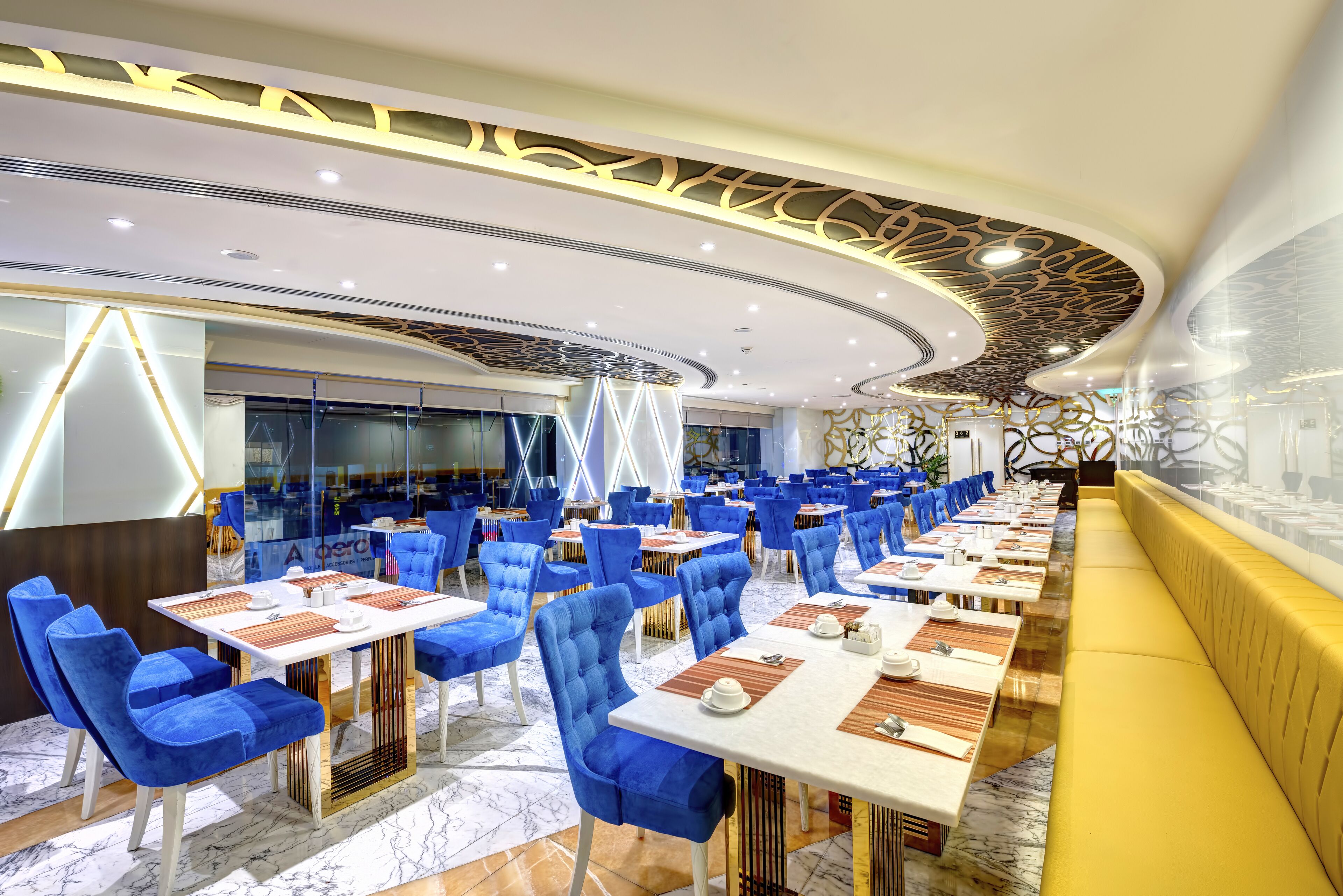 A modern restaurant with blue upholstered chairs, white tables, and a yellow bench.