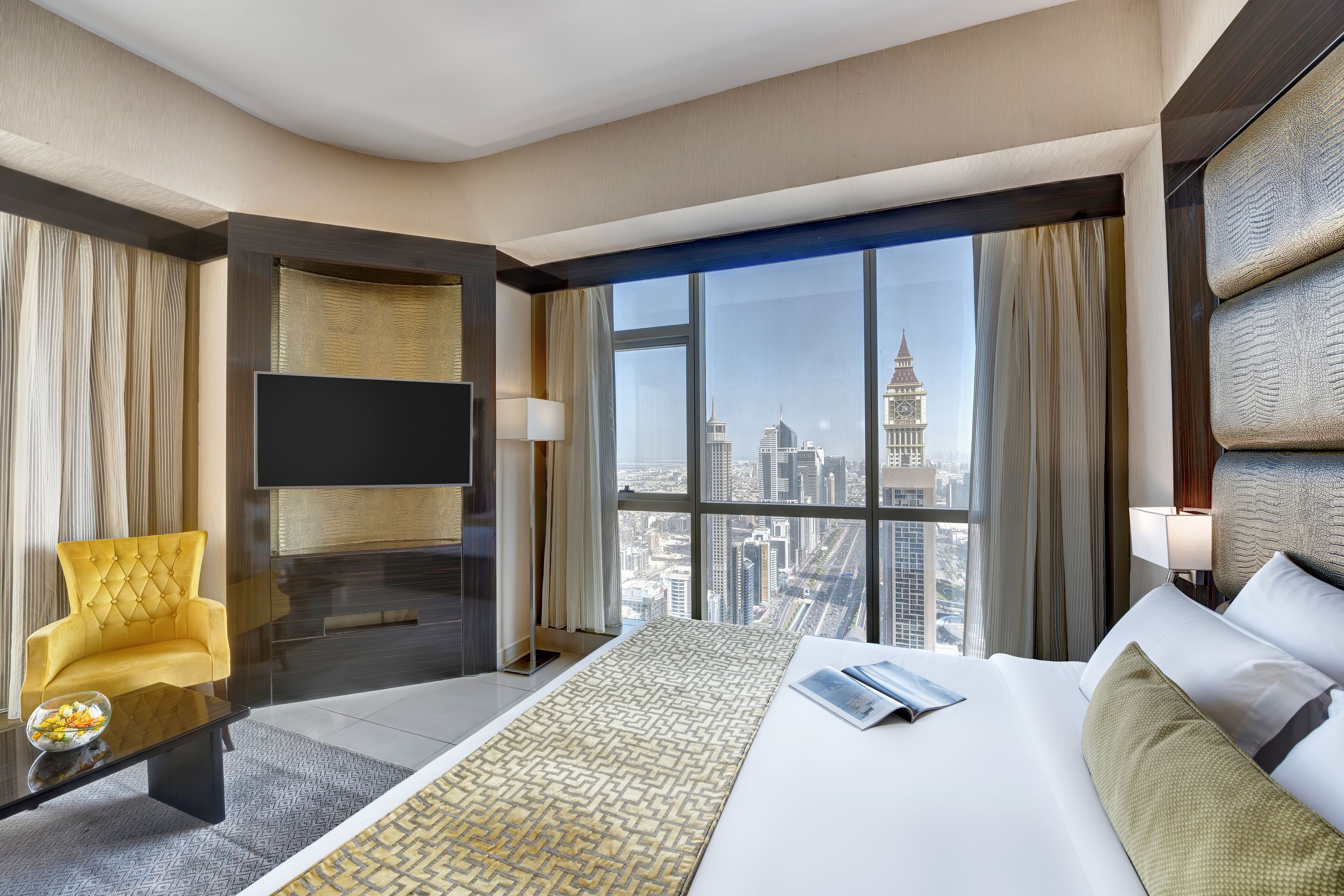 A hotel room with a large bed, a yellow armchair, a flat-screen TV, and a view of a cityscape through large windows.