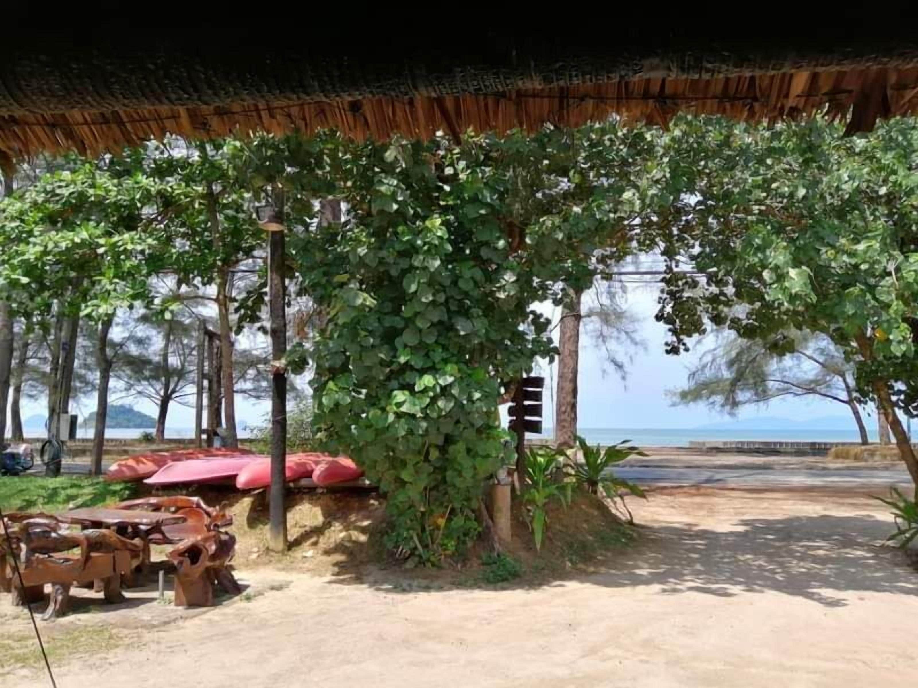 Un área de estar al aire libre con techo, mesas y sillas, rodeada de vegetación y vistas al mar.