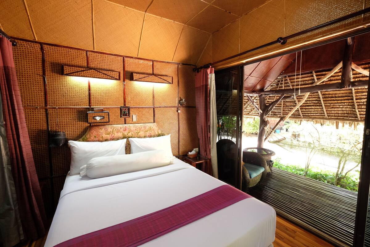 A bedroom with a bed, a nightstand, a window with curtains, and a view of an outdoor area.