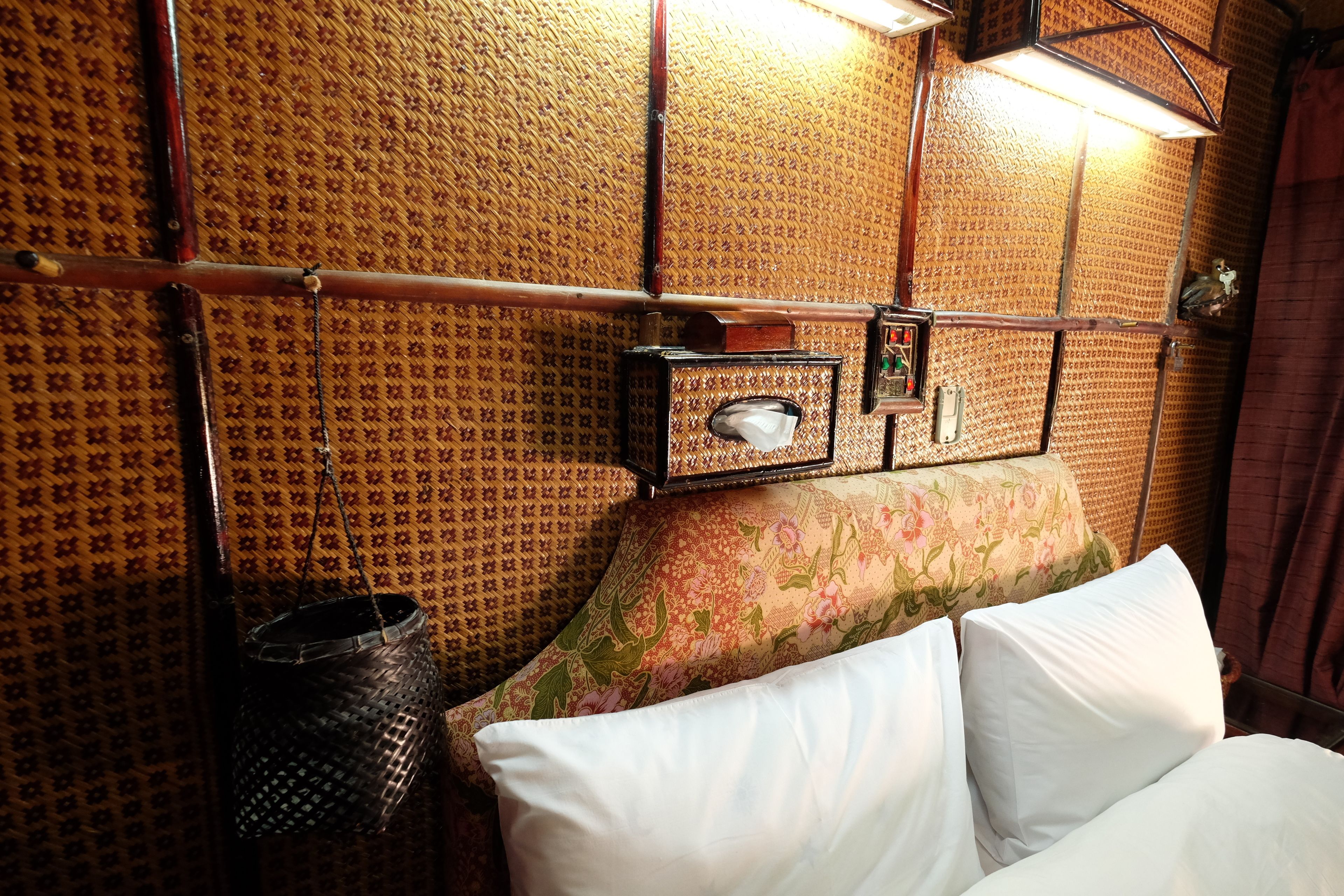 Una habitación con papel tapiz estampado, una cama con almohadillas blancas, un pequeño armario de madera y una cesta colgante.