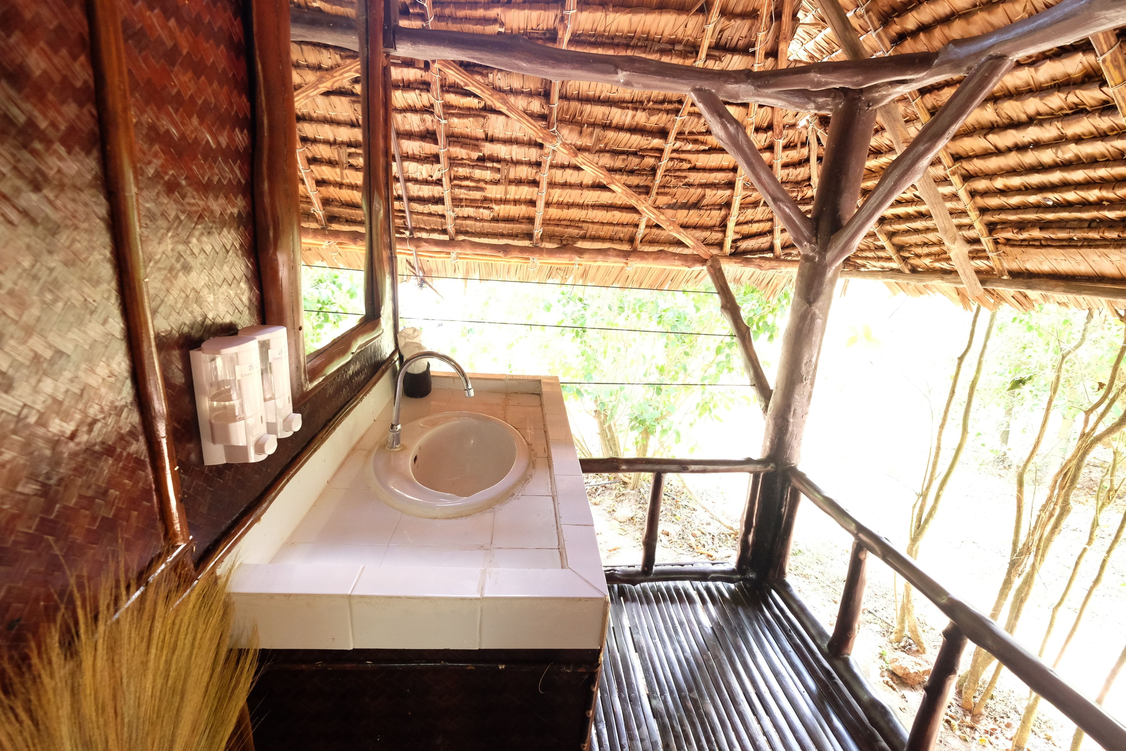 Une salle de bain rustique avec un toit de chaume, un lavabo blanc et un banc en bois.