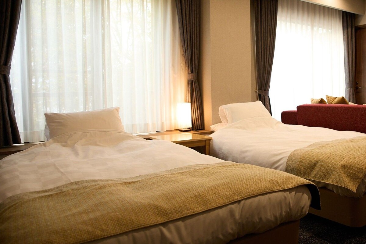 A hotel room with two beds, a nightstand, a red sofa, and a window with curtains.