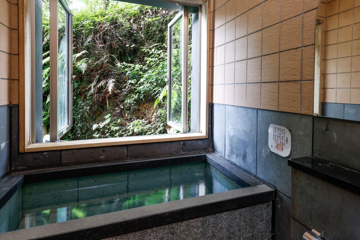 A hot spring bath with a window view of dense foliage.