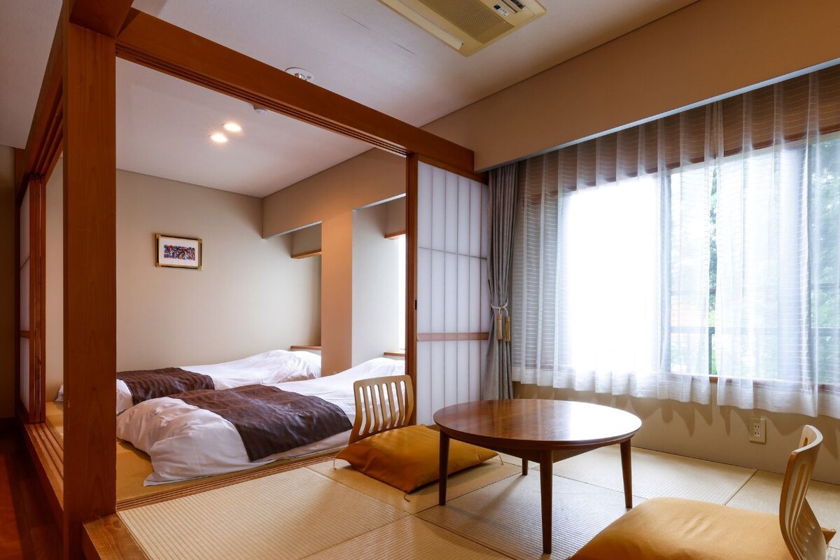A traditional Japanese-style room with tatami flooring, a low table, two beds, and a large window with sheer curtains.