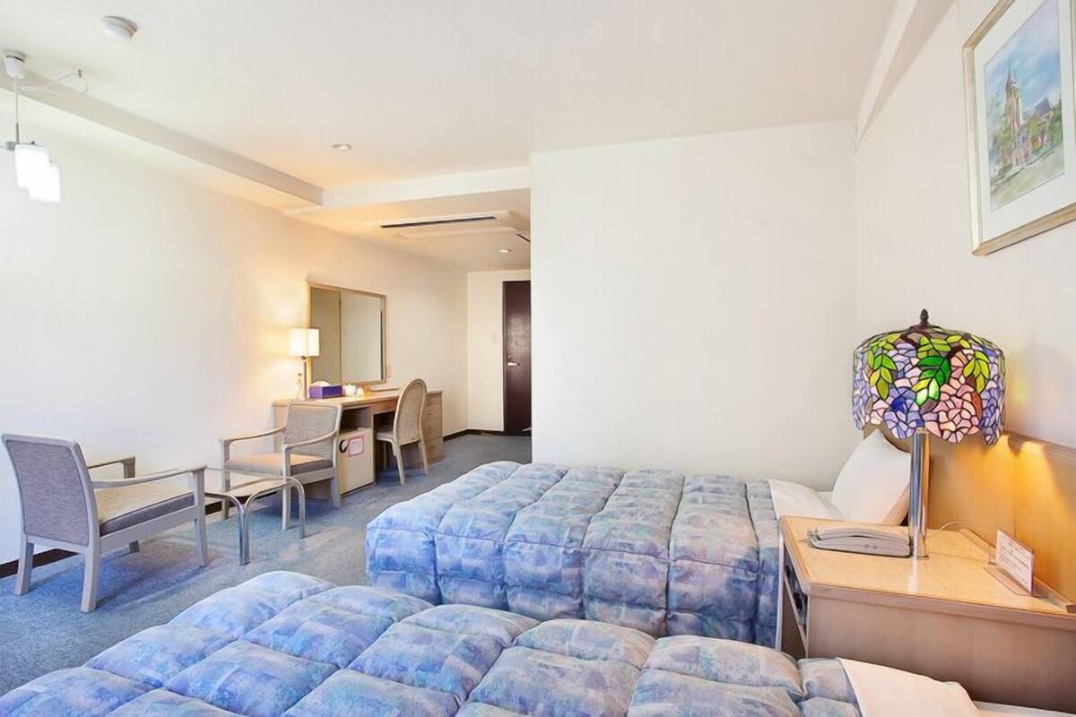 A hotel room with a bed, desk, chair, and a colorful lamp.