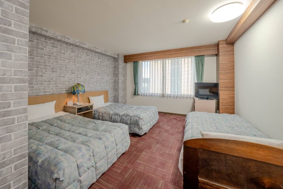 A hotel room with two beds, a brick wall, a wooden headboard, a TV, and a window with curtains.