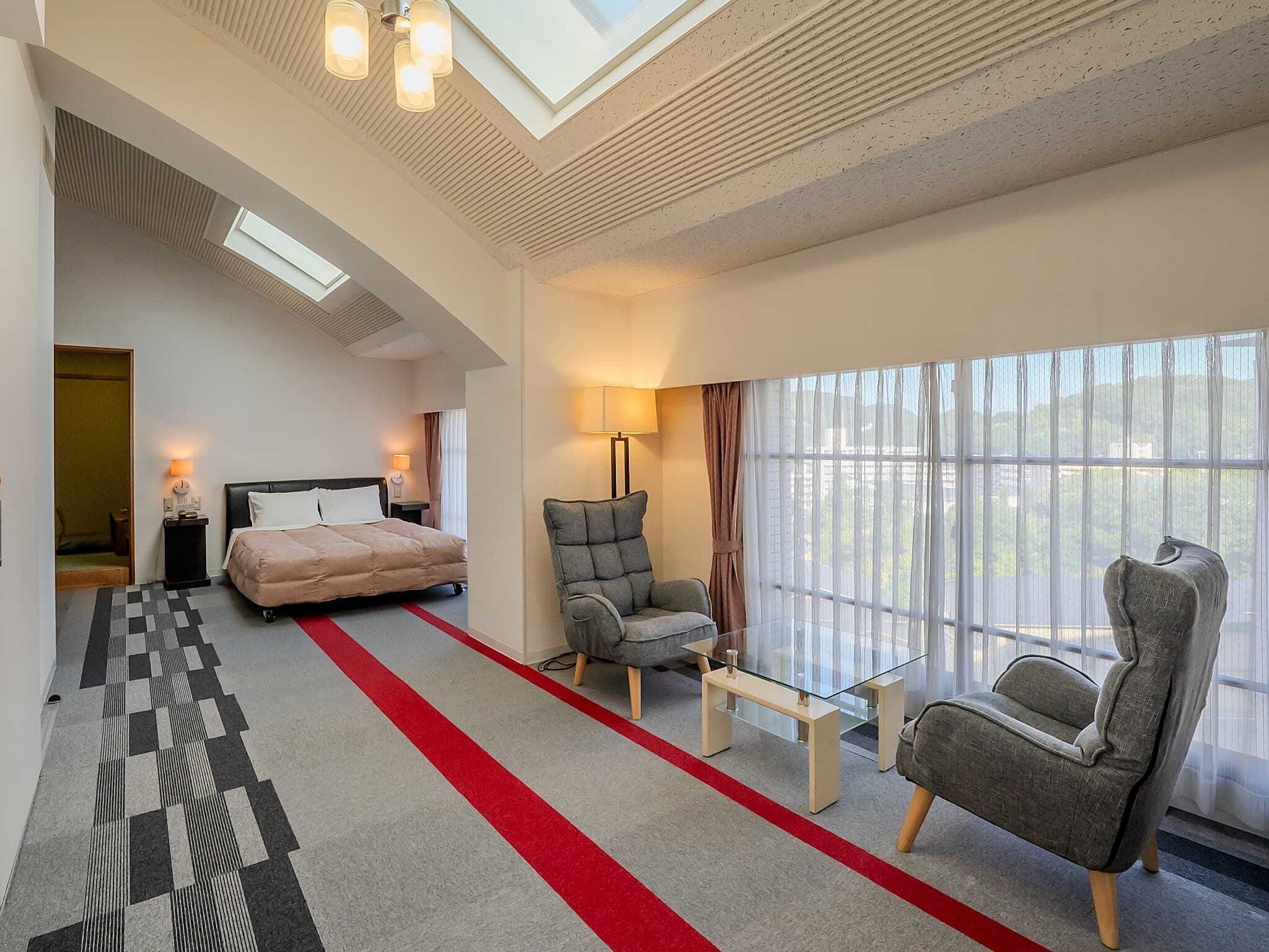 A modern hotel room with a bed, two armchairs, a glass coffee table, a lamp, and a large window with a view.