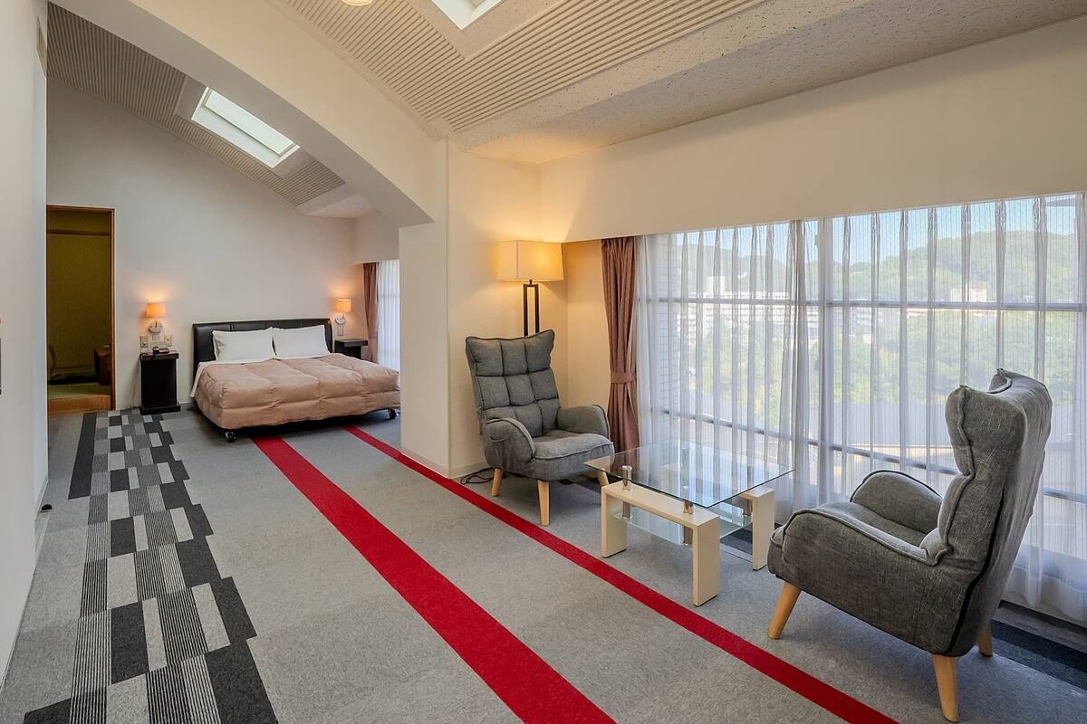 A modern hotel room with a bed, two armchairs, a glass coffee table, a lamp, and a large window with a view.