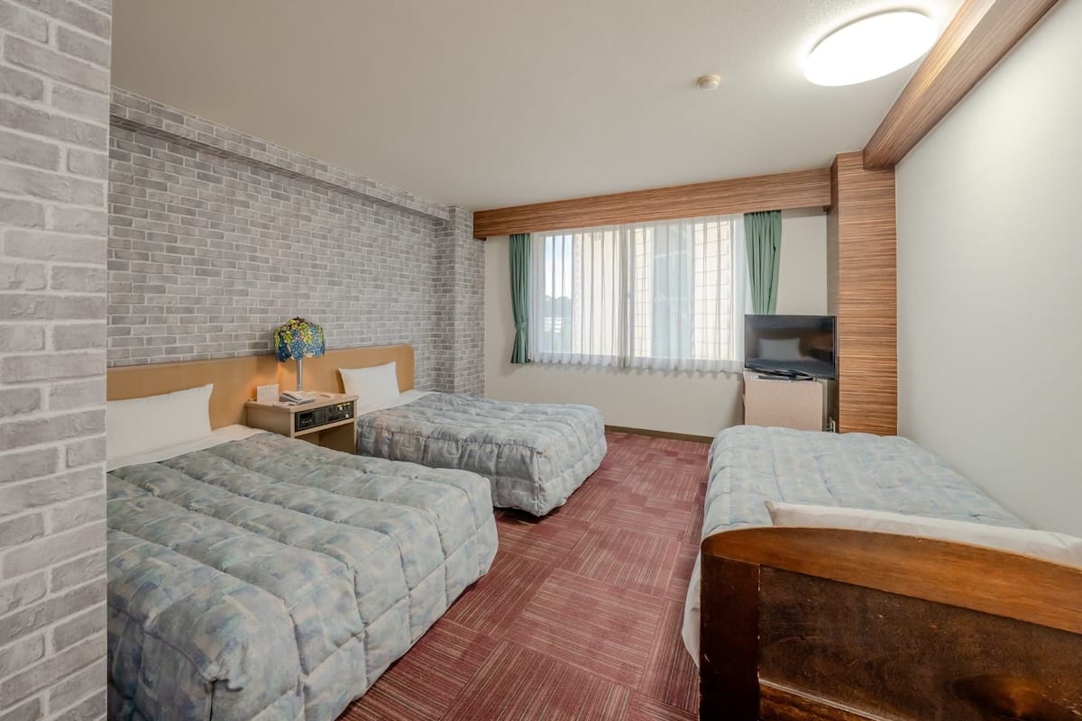 A hotel room with two beds, a brick wall, a wooden headboard, a TV, and a window with curtains.
