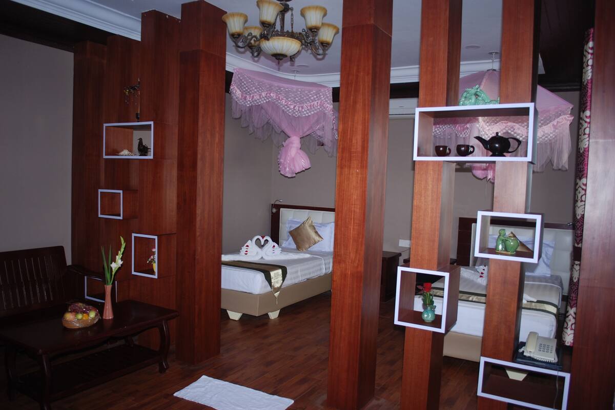 A hotel room with a wooden floor, a bed with a white headboard, a bench, a small table with a vase and flowers, and a chandelier.