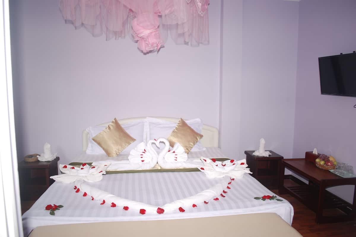 A neatly made bed with a swan pillow arrangement, a bedside table with a fruit basket, and a television mounted on the wall.