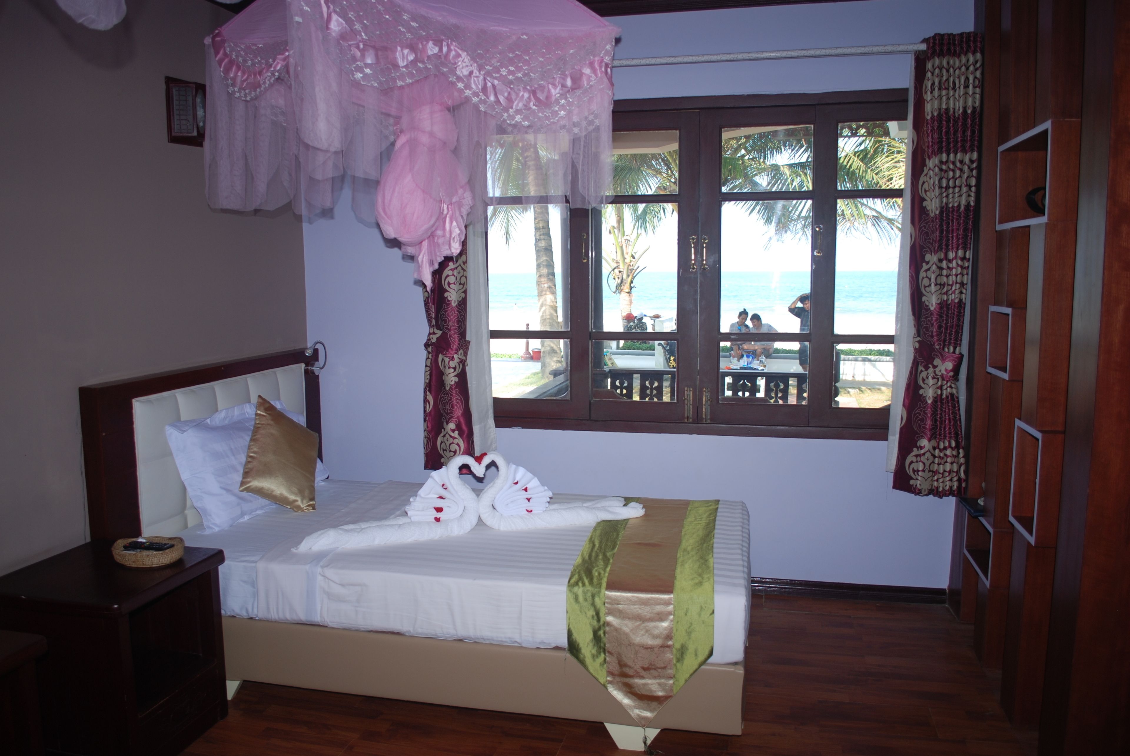 Un dormitorio con cama, mesita de noche, ventana con vista a la playa y cortina.