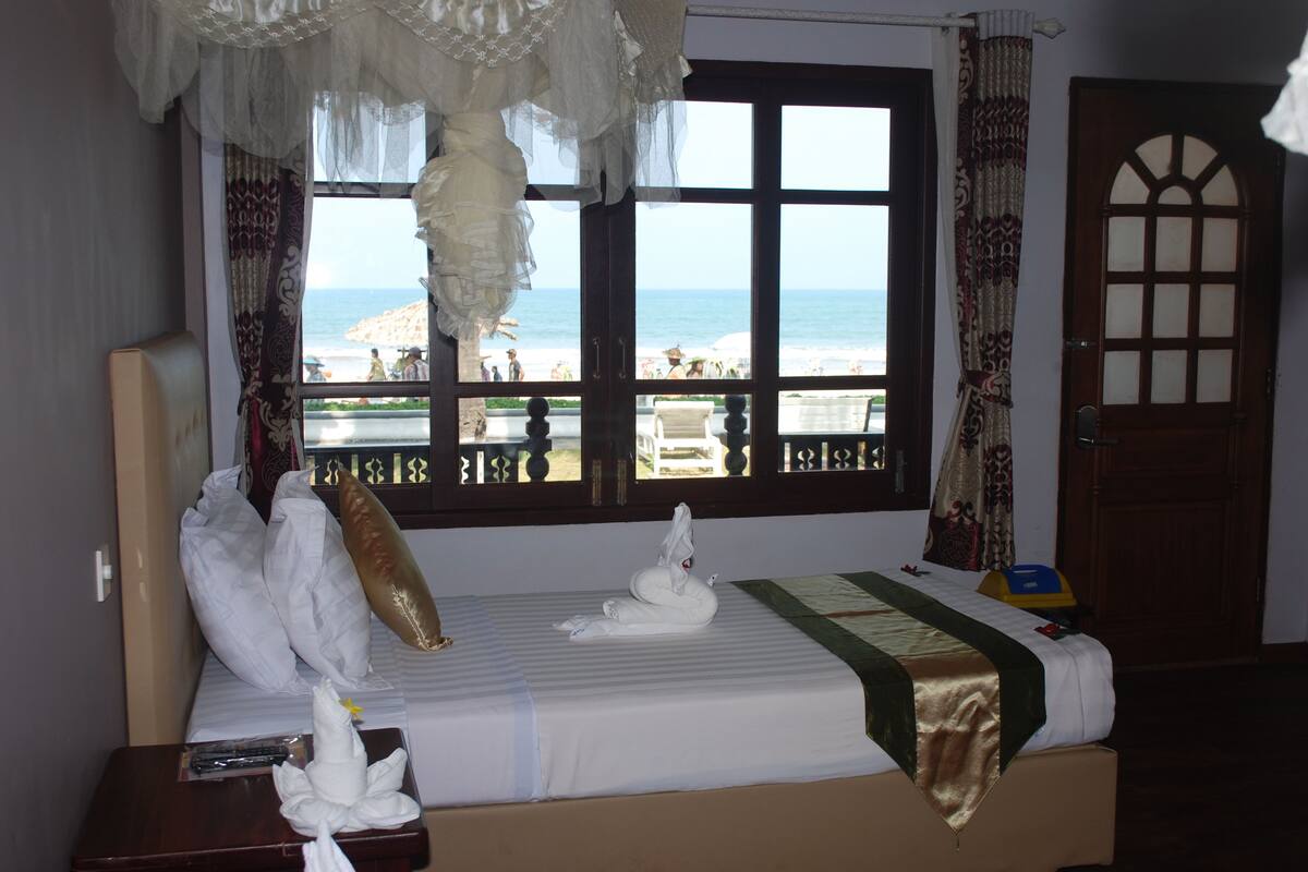 A hotel room with a bed, a window with a view of the beach, and a door.