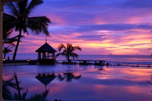 Ett semesterhotell vid stranden med en pool som speglar palmerna och en lusthus.
